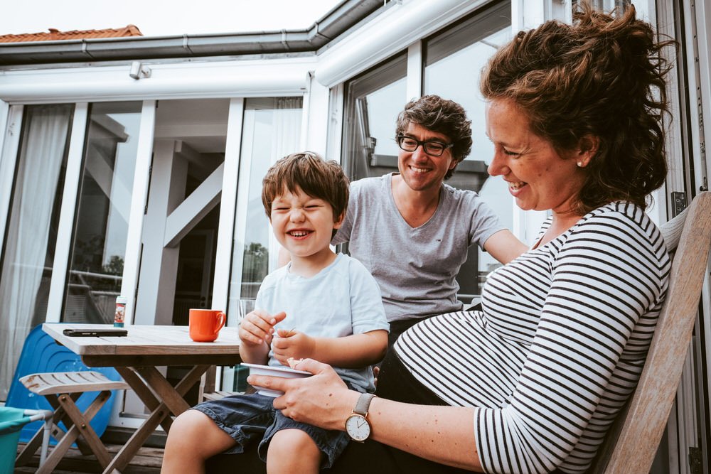 Eine lachende Frau mit lockigem Haar sitzt auf einem Stuhl, während ein junger Junge auf ihrem Schoß sitzt und lacht. Ein Mann steht neben ihnen, lächelnd und mit Arm um die Frau. Sie befinden sich auf einer Terrasse.