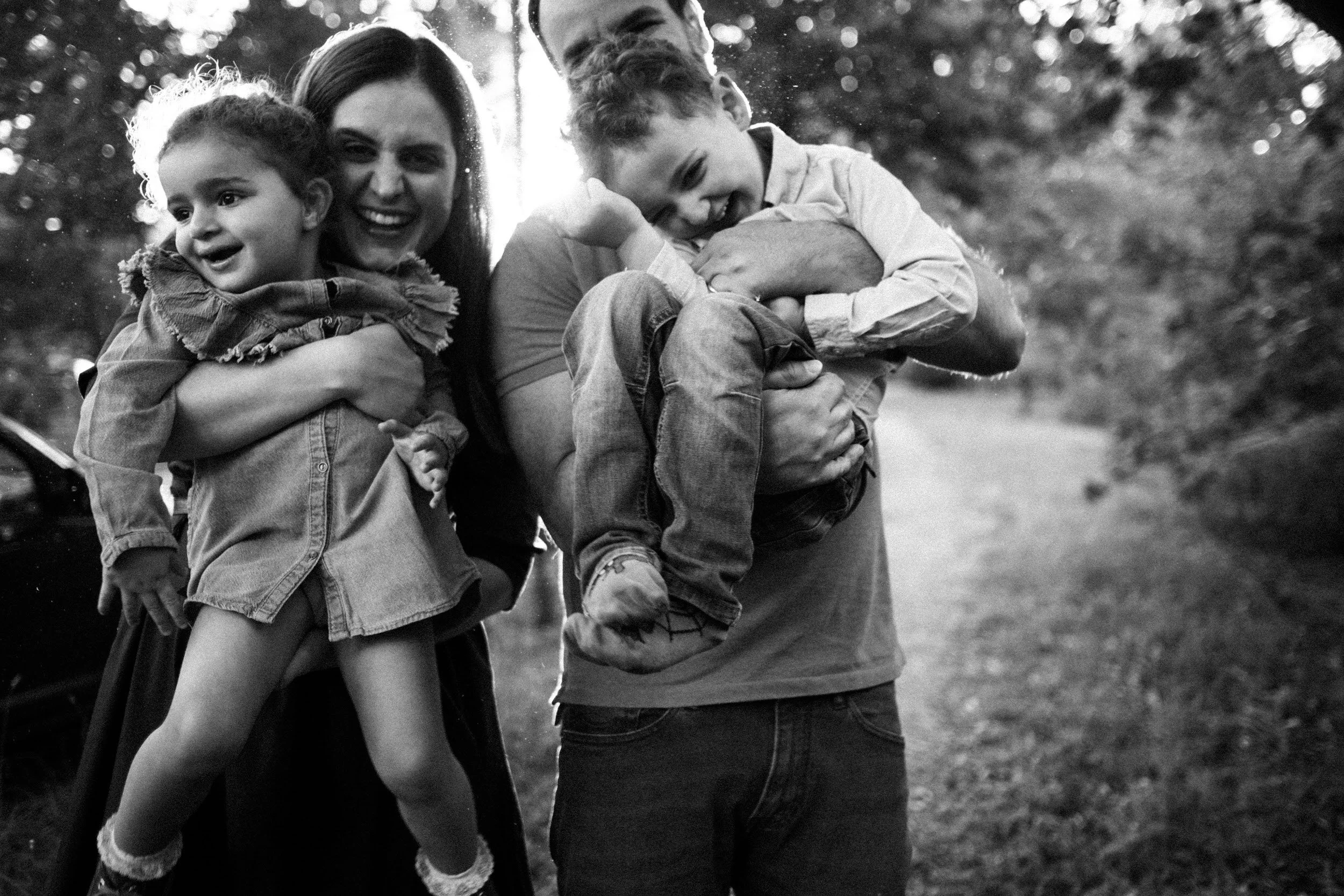 Diese emotionaler Augenblick währen eines Familien Portraits zeigt zwei Elter die ihre lachenden Kinder auf dem Arm tragen und alle den Moment zu genießen scheinen. Die warme scheinende voller Blütenstaub sieht magisch aus.