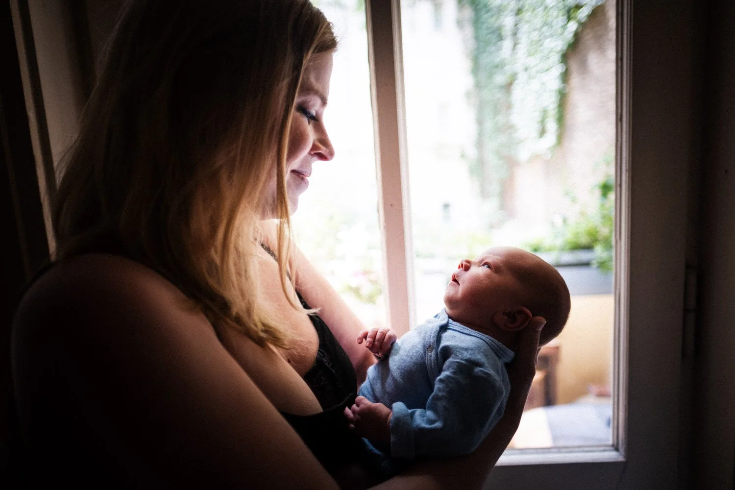 Eine Mutter hält ihr Baby vor einem Fenster, beide schauen sich liebevoll an.