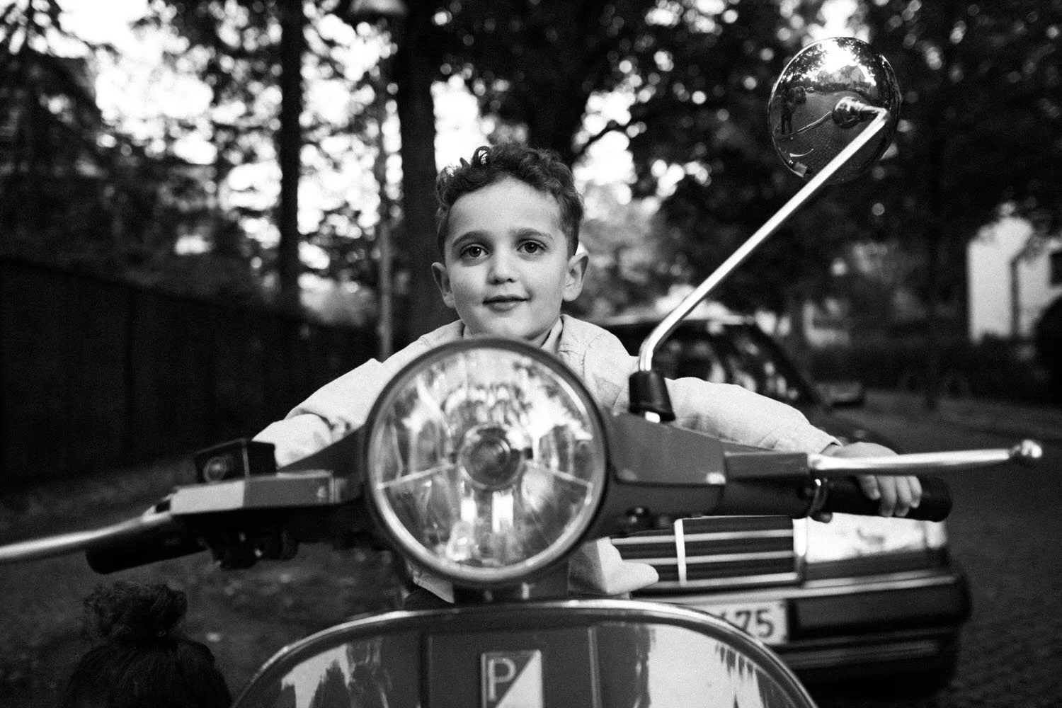 Ein Junge sitzt in der Schwarzweiß-Aufnahme auf einem Roller und lächelt in die Kamera, im Hintergrund sind Bäume und ein Auto sichtbar.
