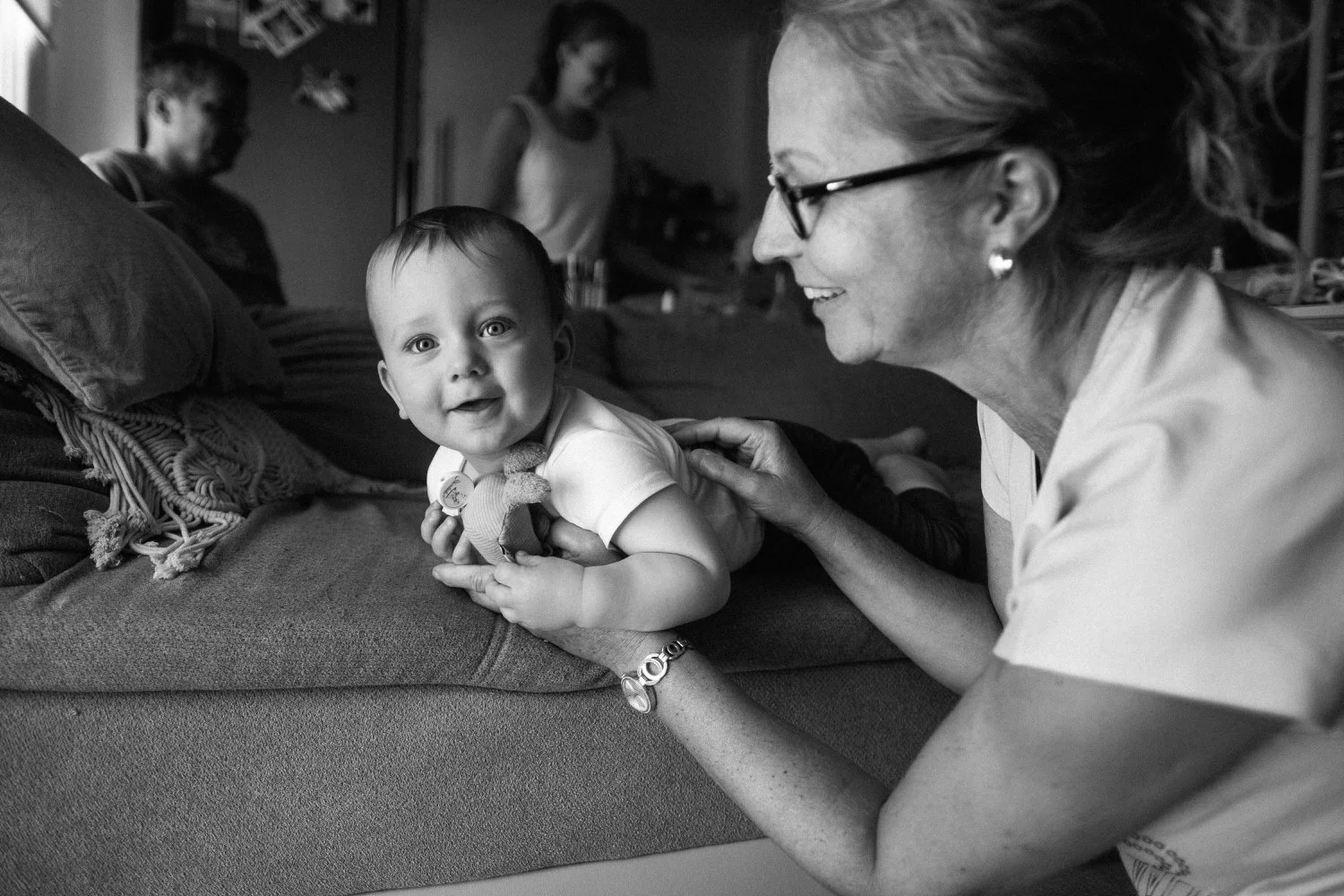 Eine Momentaufnahme in Schwarz-Weiß, in der ein Lächelndes Baby auf einem Sofa liegt, gehalten von einer älteren Frau mit Brille, während andere Personen im Hintergrund im Raum sind.