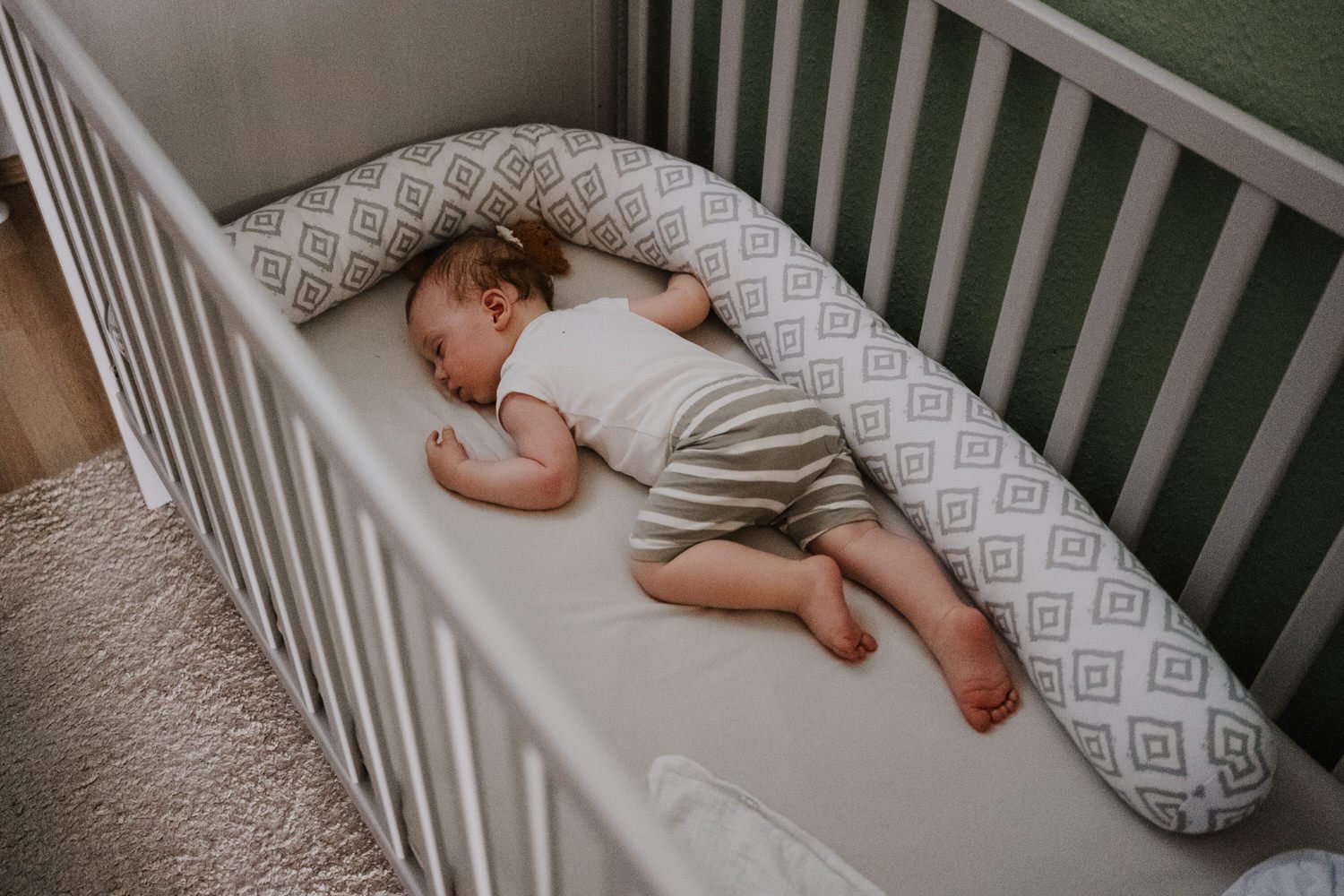 Kleines Kind schläft in einem Gitterbett auf einer Matratze mit einem Nackenrollen-Kissen.