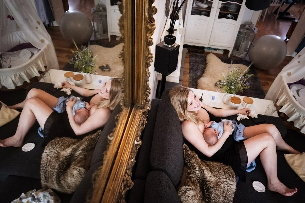 Szene aus einer Homestorie: Eine junge Mutter sitzt auf einem schwarzen Sofa, stillt ihr Baby und lächelt glücklich. Das Bild wird durch einen großen, goldgerahmten Spiegel reflektiert, der die Szene zeigt.