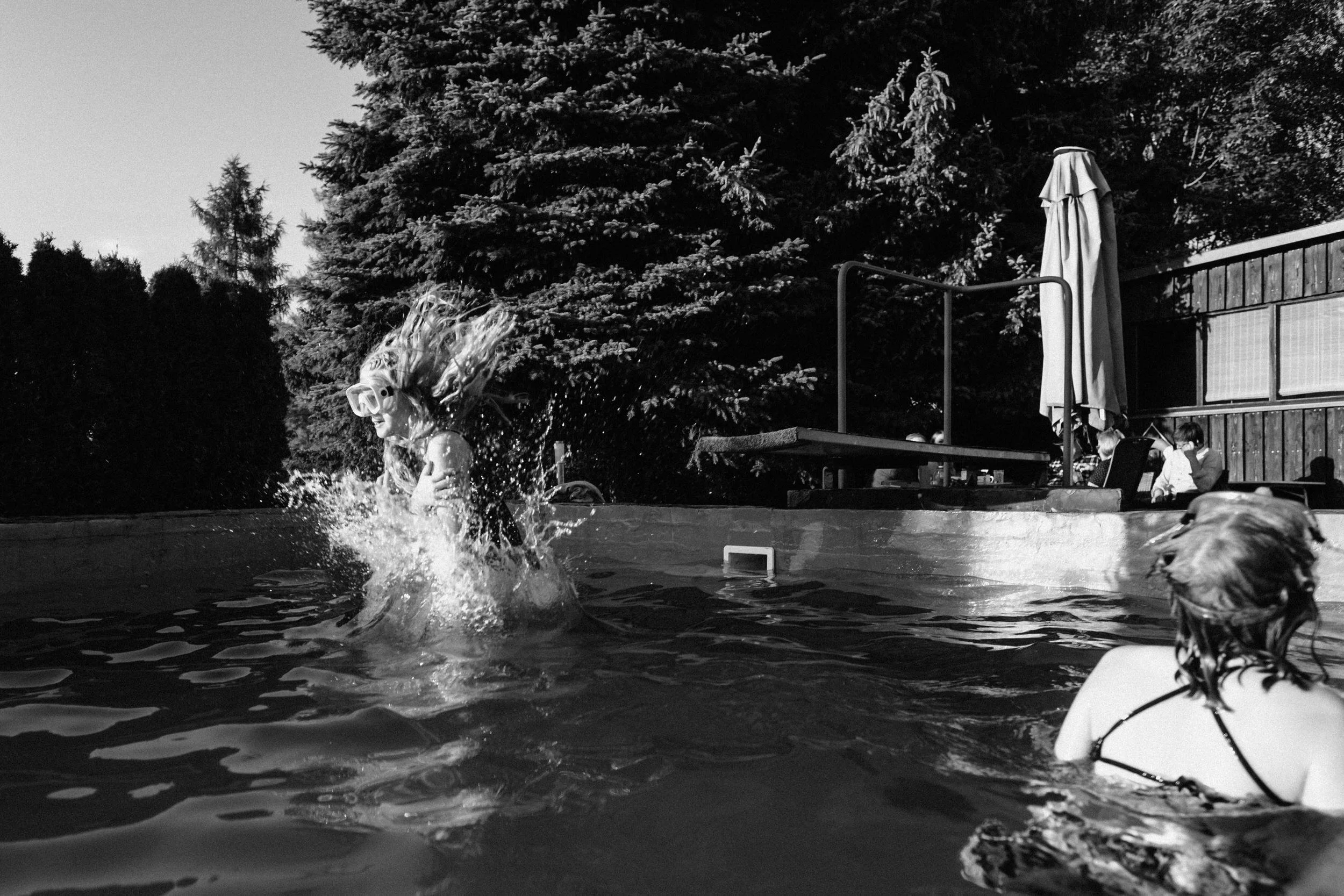 Diese schwarz-weiße Momentaufnahme während eines Familienfotoshootings zeigt einen dynamischen Sprung in das Becken eines Pools von einem Sprungbrett. Dem Mädchen mit Taucherbrille fliegen die langen Haare in alle Richtungen.