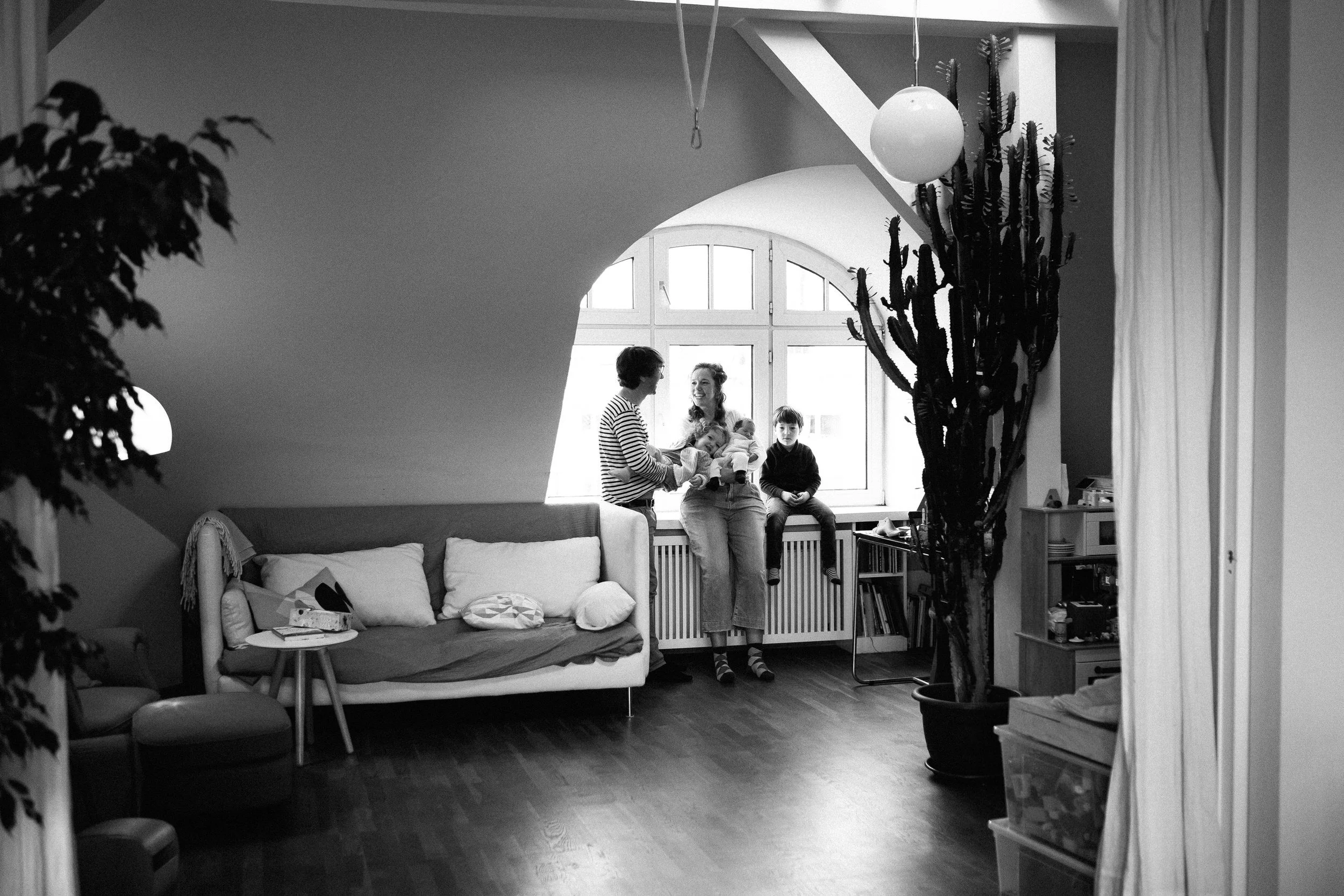 Eine weite Aufnahme eines Dachgeschosswohnung in Berlin in Schwarz-Weiß. Im Hintergrund sieht man eine Familie glücklich am Fenster stehend. Im Vordergrund ist eine stilvolle Einrichtung mit Planzen.