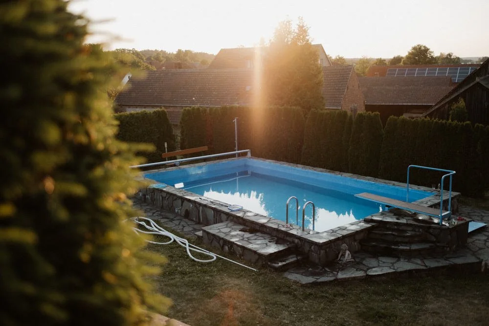 Ein Swimmingpool in einem Garten bei Sonnenuntergang, umgeben von Hecken und Häusern mit Solaranlagen auf den Dächern.