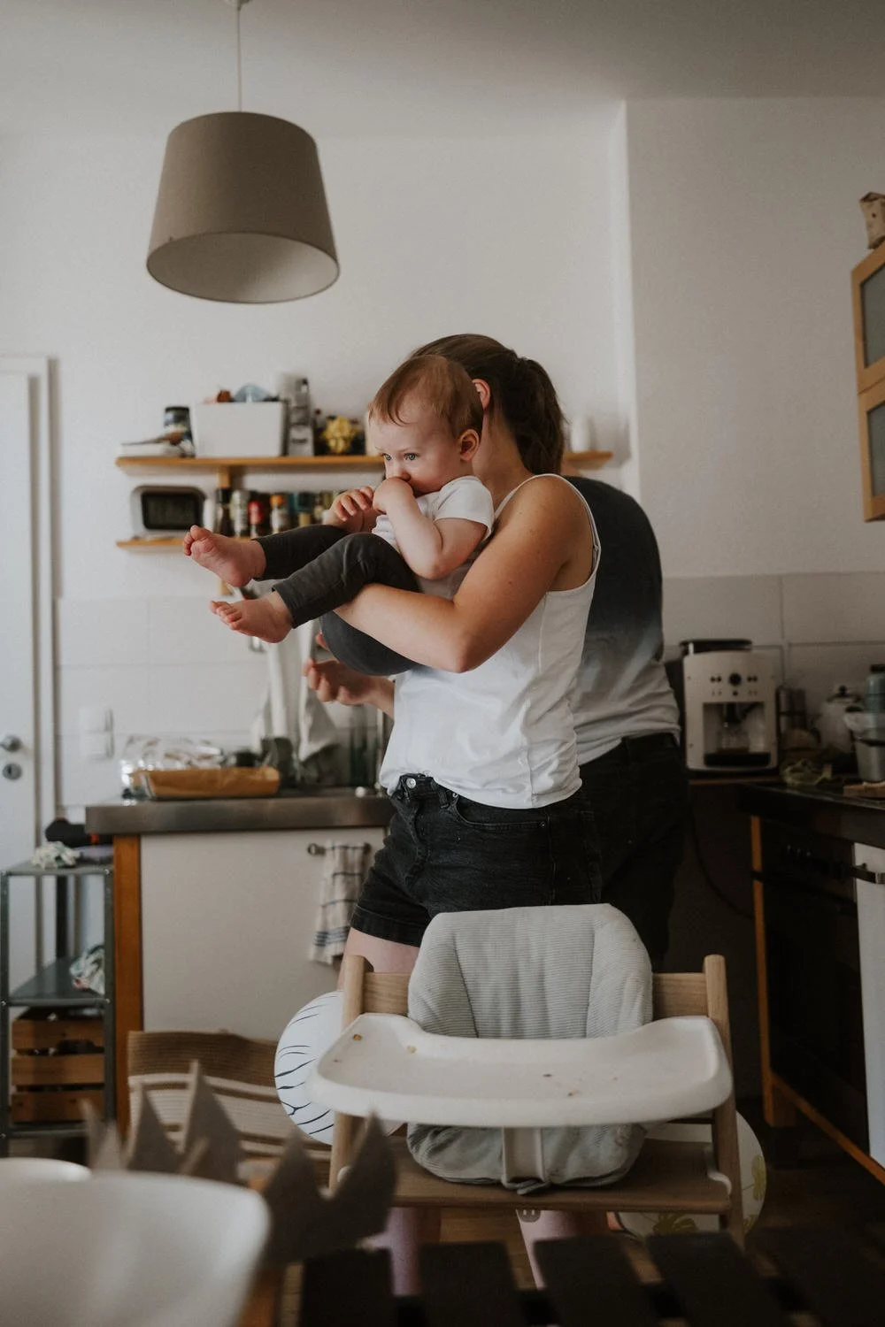 Eine Frau hält ein Baby in der Küche, im Hintergrund sind helle Küchenschränke und kleine Geräte sichtbar.