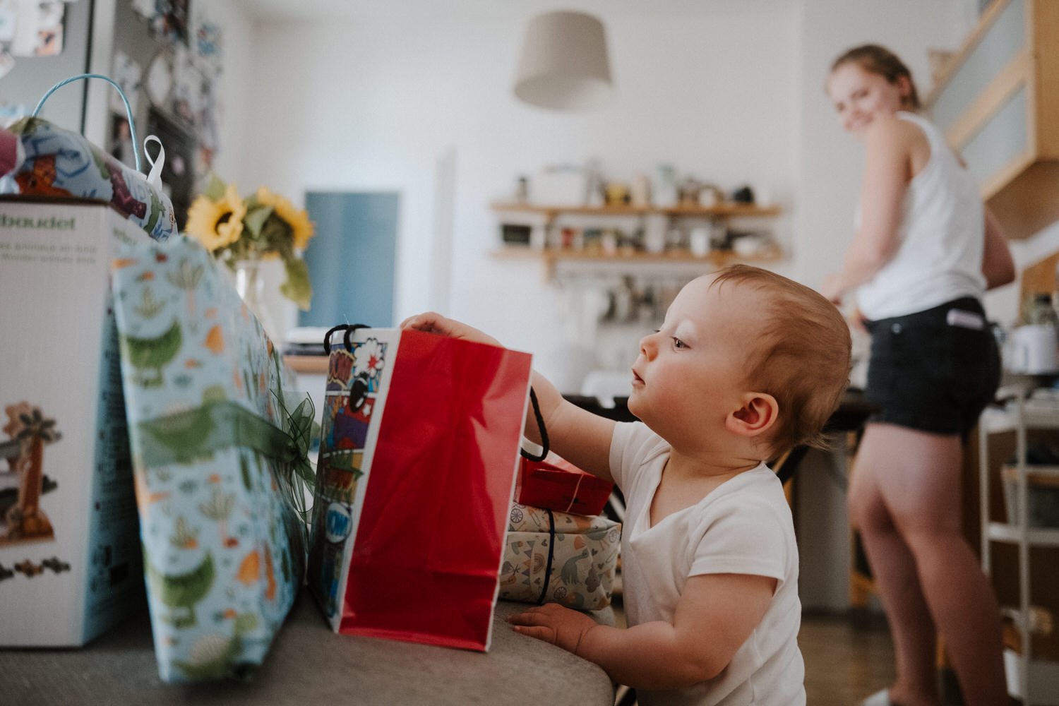Ein Baby steckt seine Hand in eine Geschenkverpackung auf einem Tisch in der Küche, während eine junge Frau im Hintergrund arbeitet.