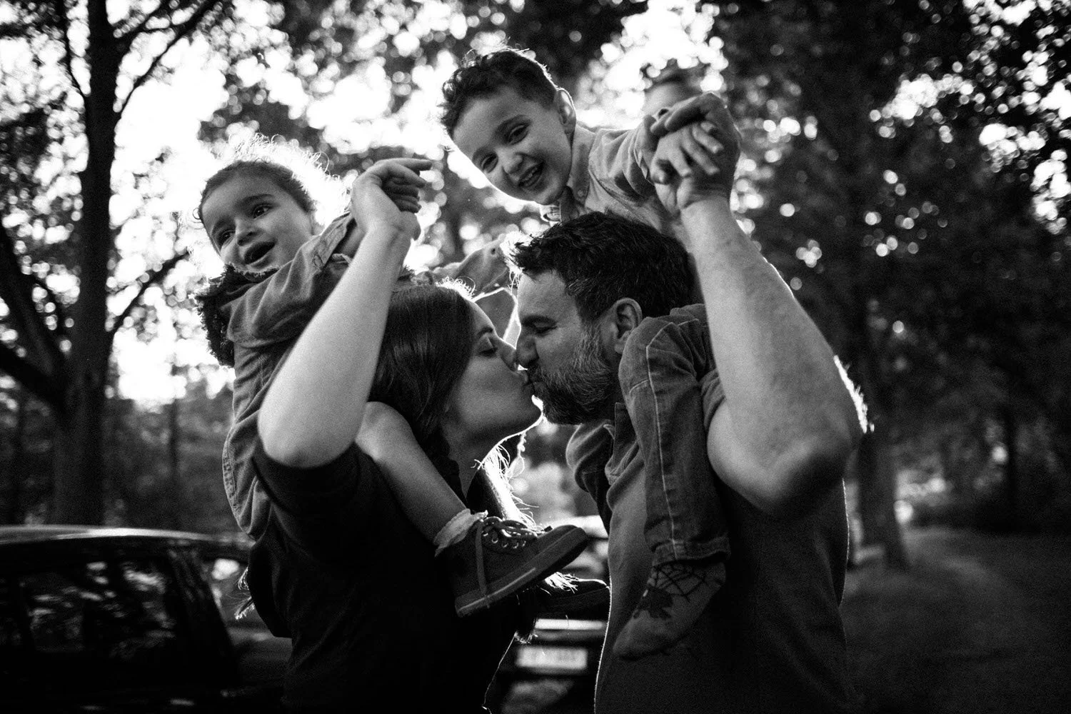 Ein glückliches Familienfoto bei Sonnenuntergang im Park, zeigt ein Paar, das sich küsst, während zwei Kindern auf den Schultern sitzen und lachen.