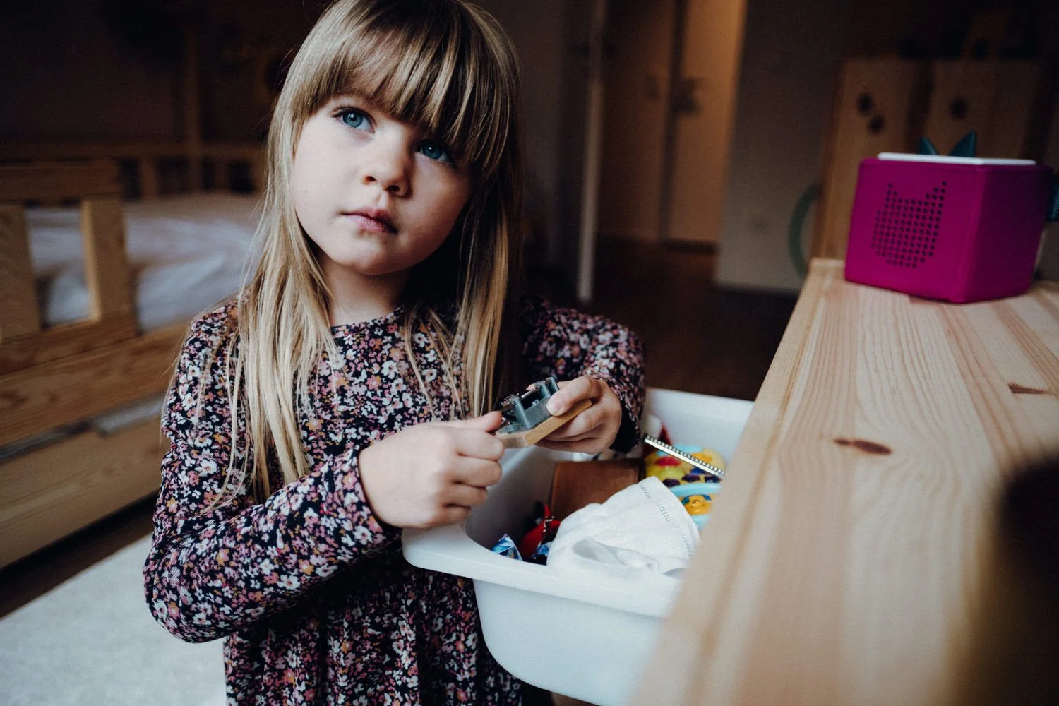 Ein Mädchen mit blonden Haaren und einem bunten Kleid schaut lauscht im Kinderzimmer ganz konzentriert auf die Spieluhr die sie hält.
