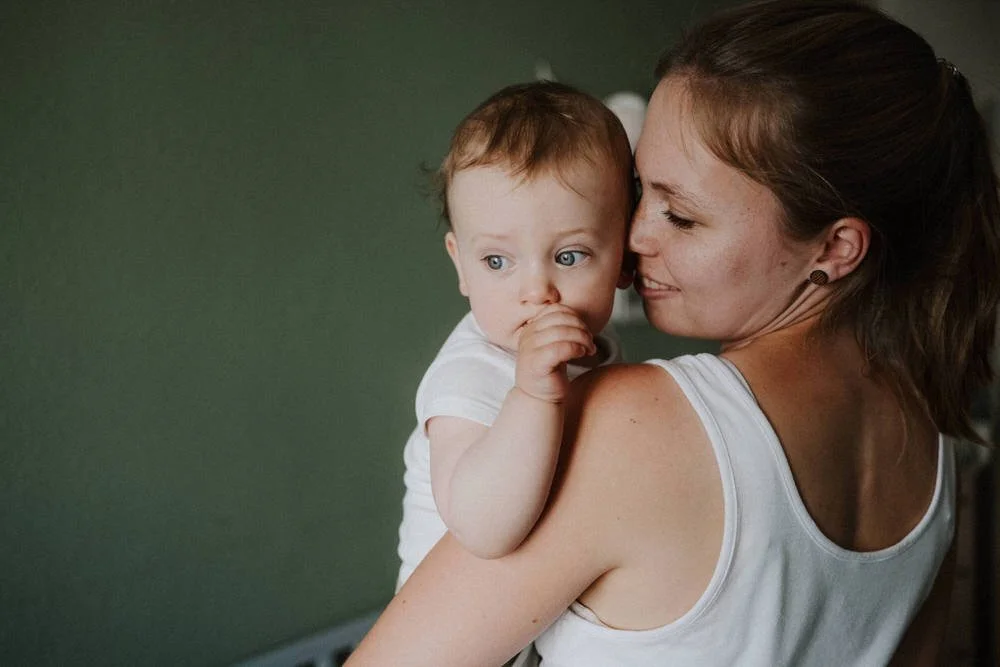Eine junge Frau hält ein Baby mit blauen Augen und braunen Haaren, das in ihren Armen liegt, vor einem grünen Hintergrund. Die Koepfe berühren sich zaertlich. Ein inniger Moment zwischen Mutter und Sohn.