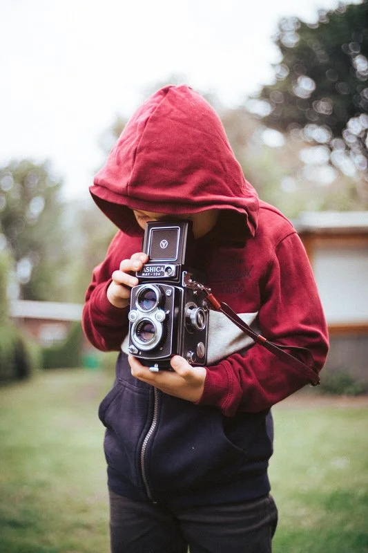 Ein Kind in rotem Hoodie hält eine alte analoge Kamera im Freien. Das Kind ist sehr konzentriert und stimmungsvoll in der grünen Natur eingefangen.