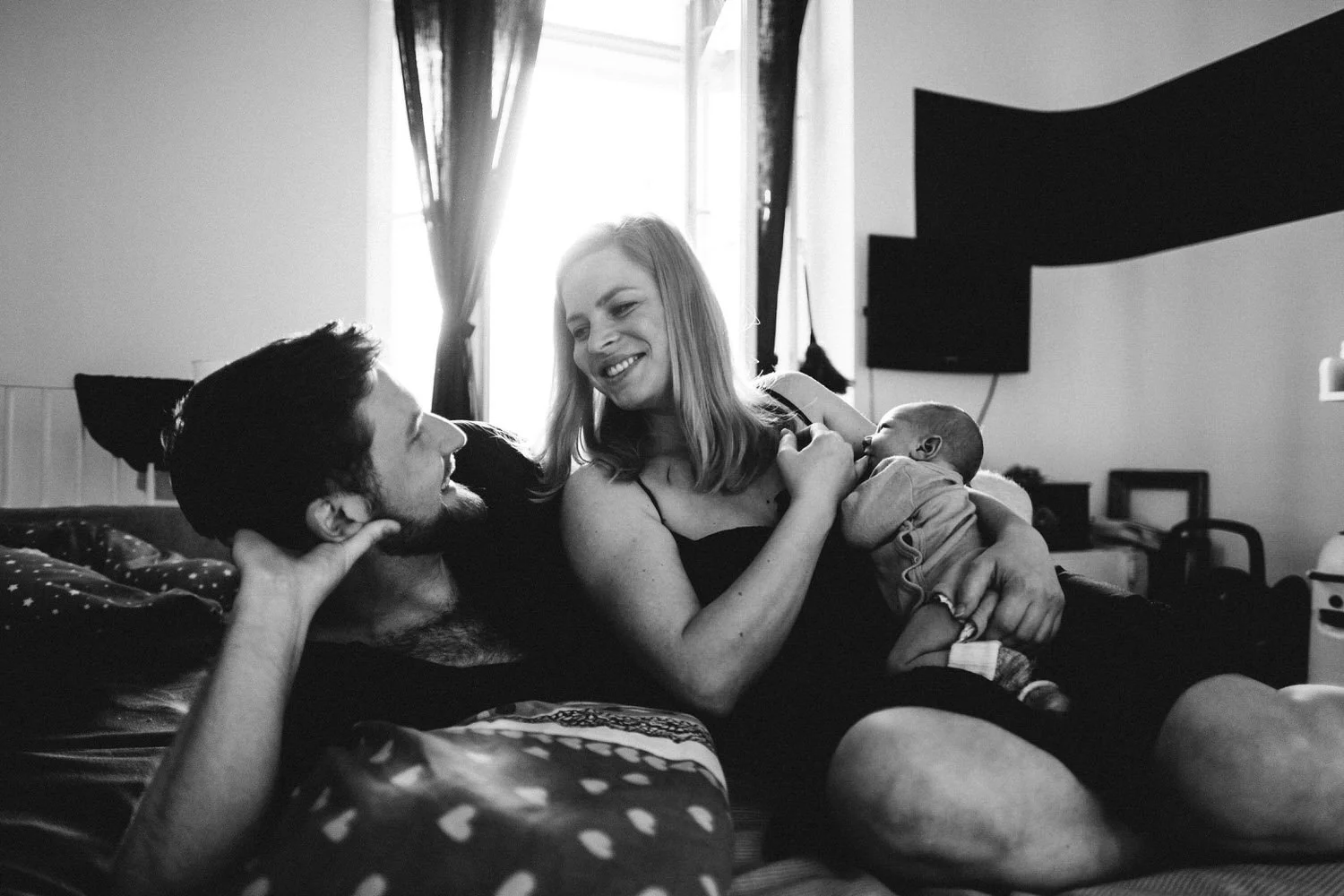 Ein Mann und eine Frau sitzen auf den Bett, die Frau hält ein kuschelndes Baby im Arm, während der Mann sie liebevoll ansieht. Die Szene ist in einem Schlafzimmer, mit einem Altbaufenster mit viel Licht.