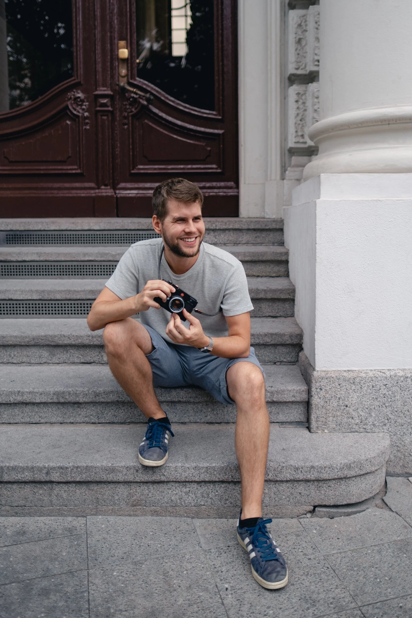 Fotograf Marvin Girbig sitzt lachend auf einer Treppe und hält seine schwarze Leica M Kamera einsatzbereit in den Händen.