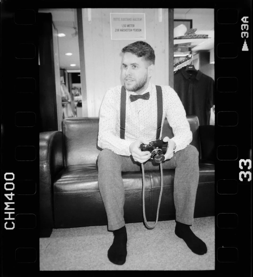 Marvin Girbig sitzt mit seiner Leica Kamera auf einem schwarzen Leder-Sofa, trägt ein helles Hemd mit Punkten, eine Fliege und Hosenträger. Der Fotograf ist hier während einer Reportage festgehalten worden.