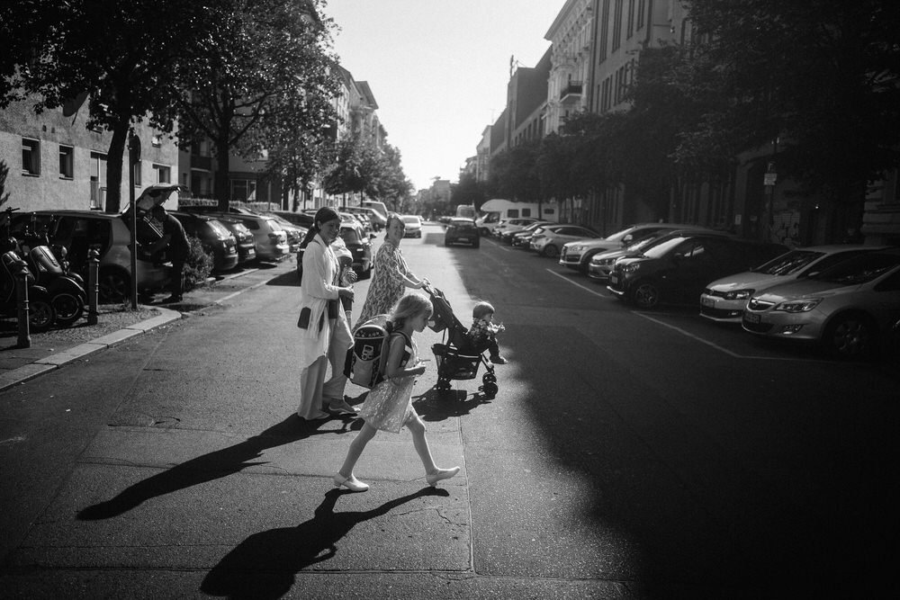 Zwei Frauen und ein Schulkind mit Rucksack und einem Kind im Kinderwagen, die die Straße überqueren, auf einer sonnigen Straße in Berlin mit parkenden Autos und Bäumen auf beiden Seiten.