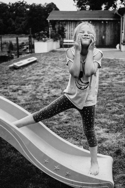 Ein junges Mädchen steht schräg auf einer Rutsche und zieht eine Grimasse mit den Händen im Gesicht - eine lustiger Momentaufnahme die die Verspieltheit des Kindes zeigt.