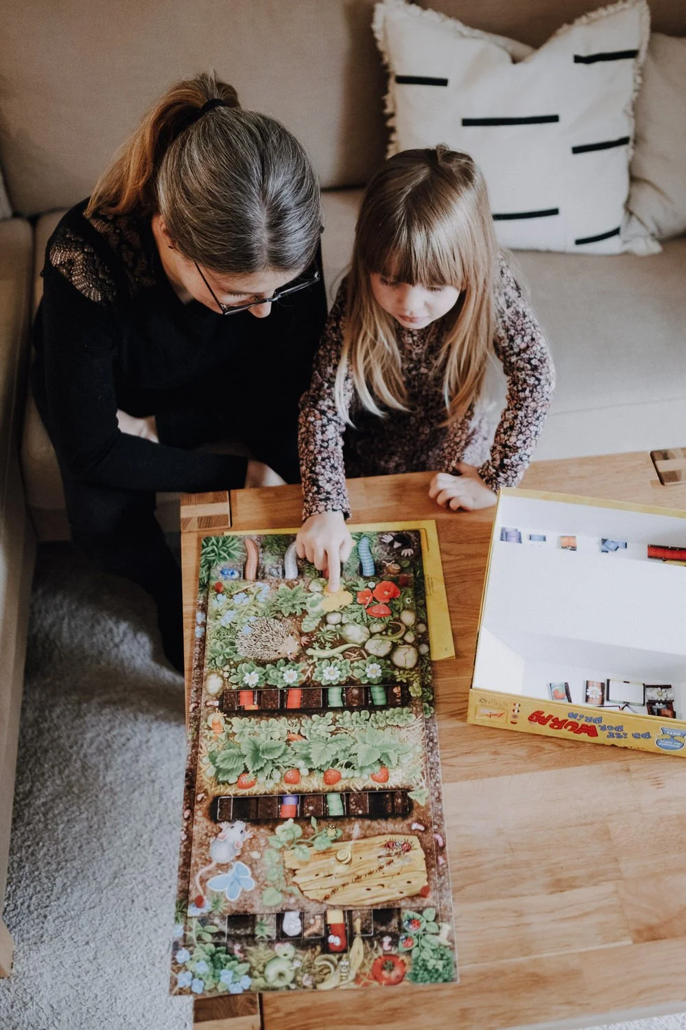 Eine Mutter und ein Kind spielen gemeinsam ein Brettspiel an einem Holztisch im Wohnzimmer. Die Aufnahme ist von oben mit Blick auf das Spielbrett angefertigt.