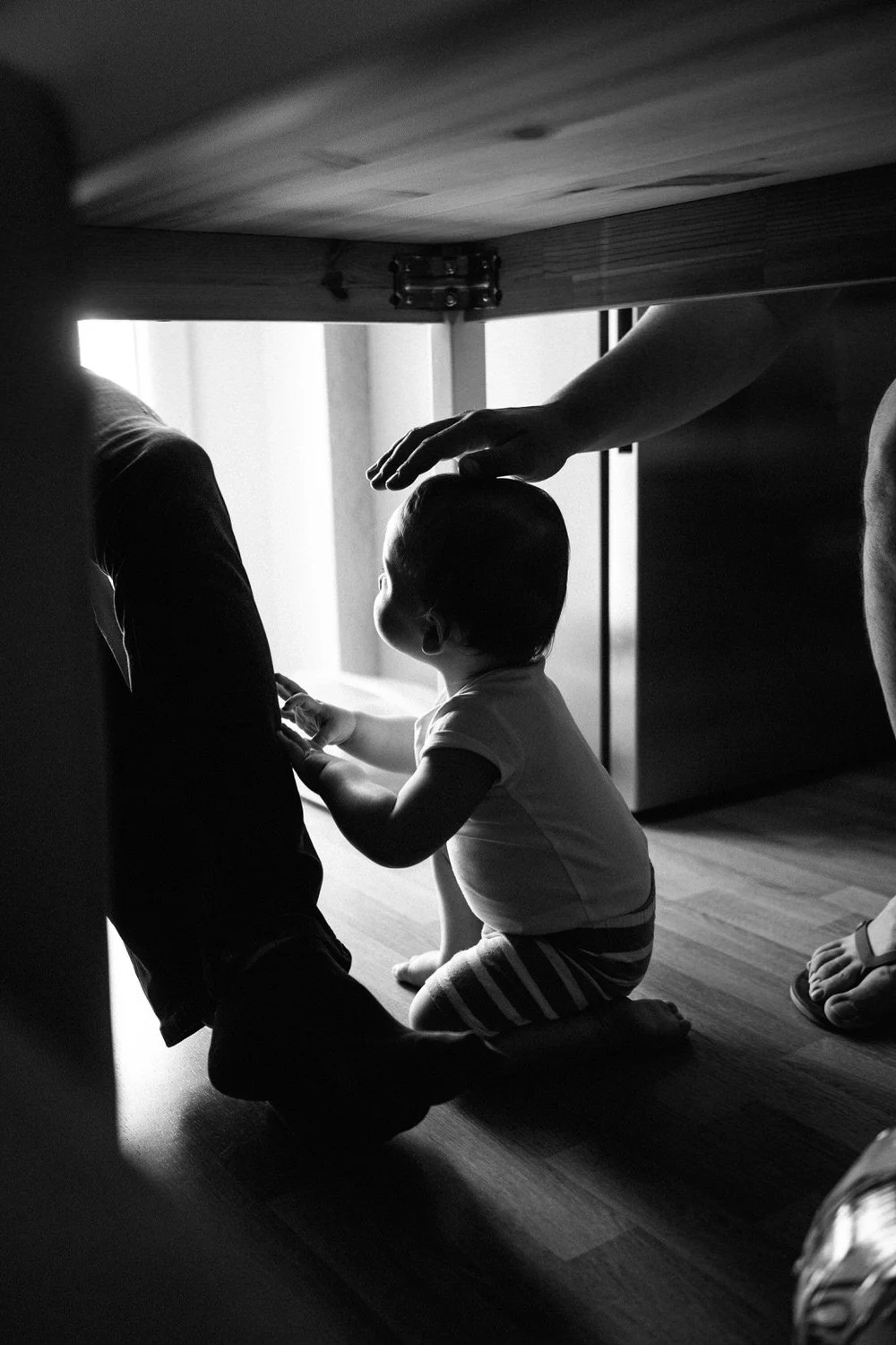 Ein Kind sitzt unter einem Tisch und berührt das Bein eines Erwachsenen und wird gestreichelt. Mit natürlichem Licht im Hintergrund im Schwarz-Weiß-Stil.