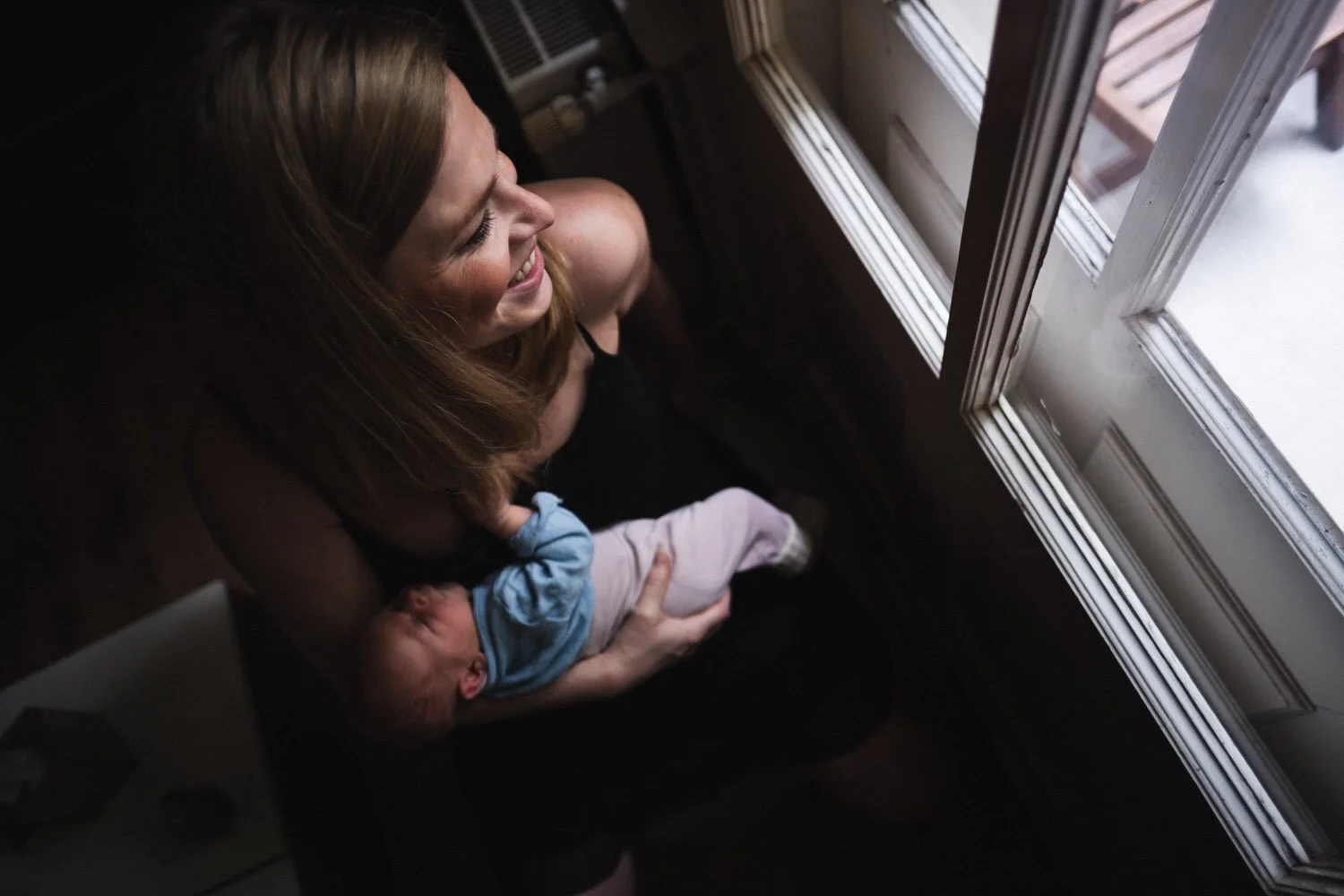 Eine lächelnde Mutter hält ein neugeborenes Baby in den Armen, während sie neben einem Fenster sitzt.