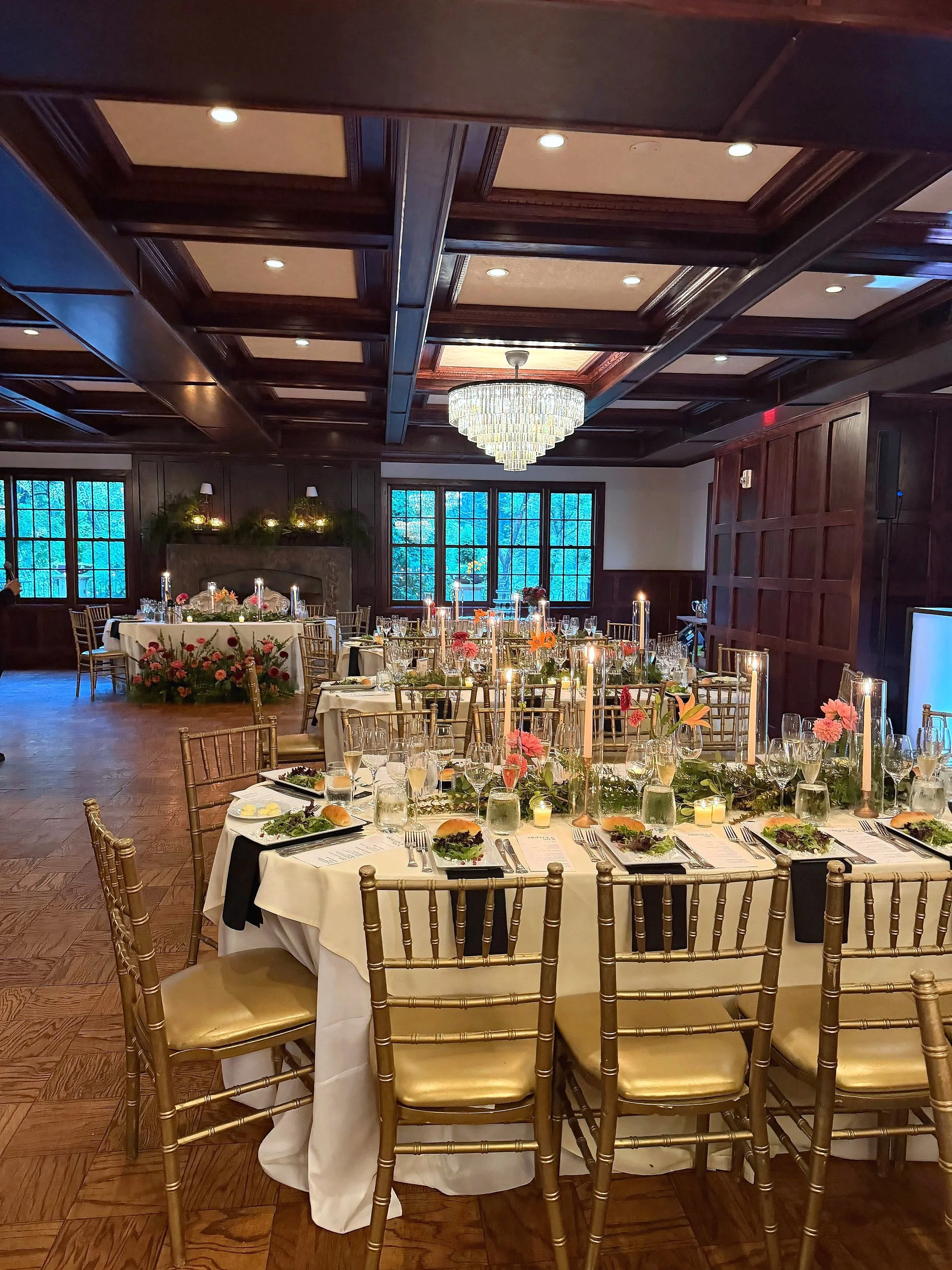 Elegant banquet hall set up for a formal dinner with round tables, floral centerpieces, candle decorations, and gold chairs. A large chandelier hangs from the ceiling, and windows in the background let in natural light.