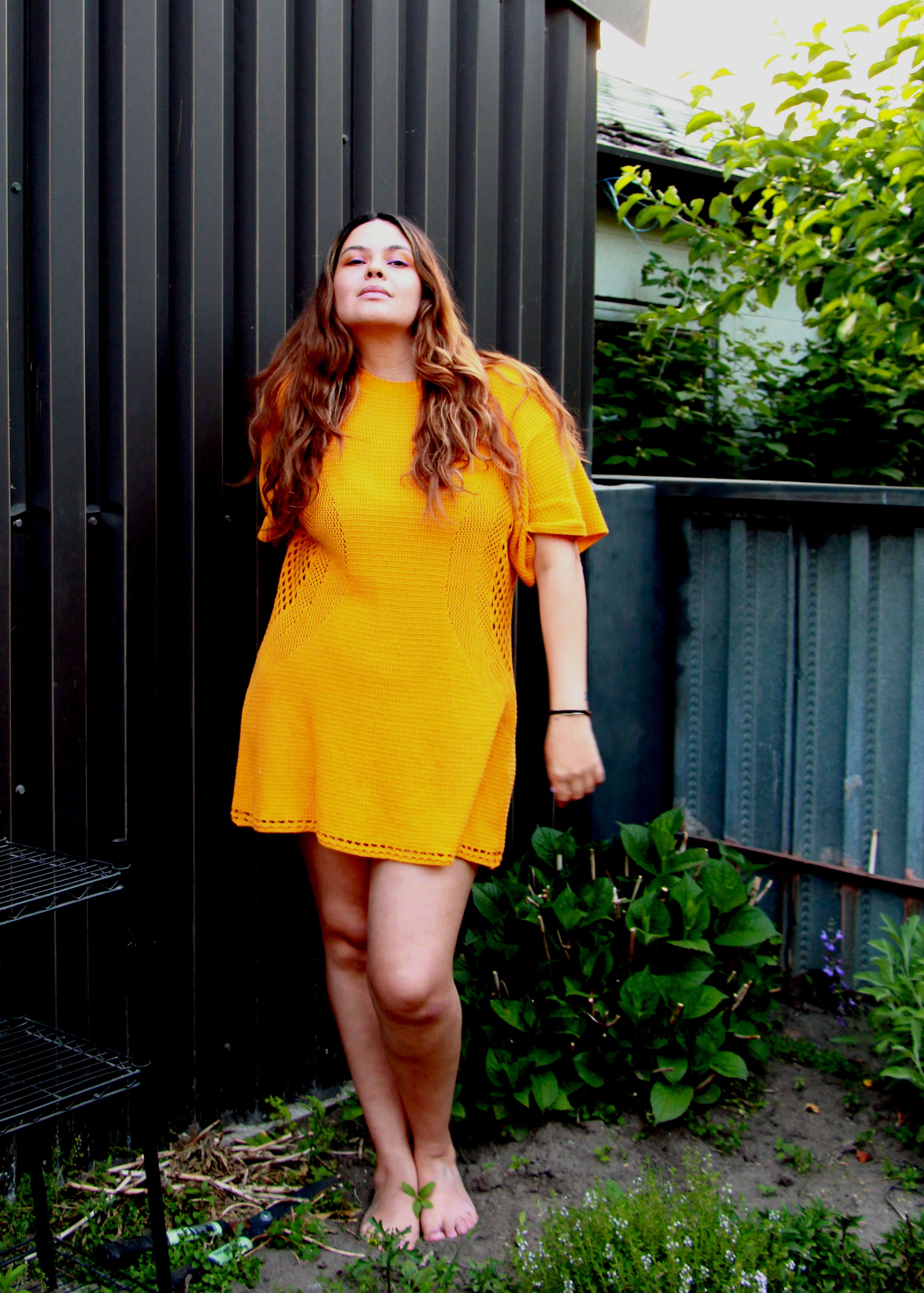  A woman with long brown hair is standing in a garden, in front of a metal shed and wood fence. She is smiling and wearing a marigold yellow dress. 