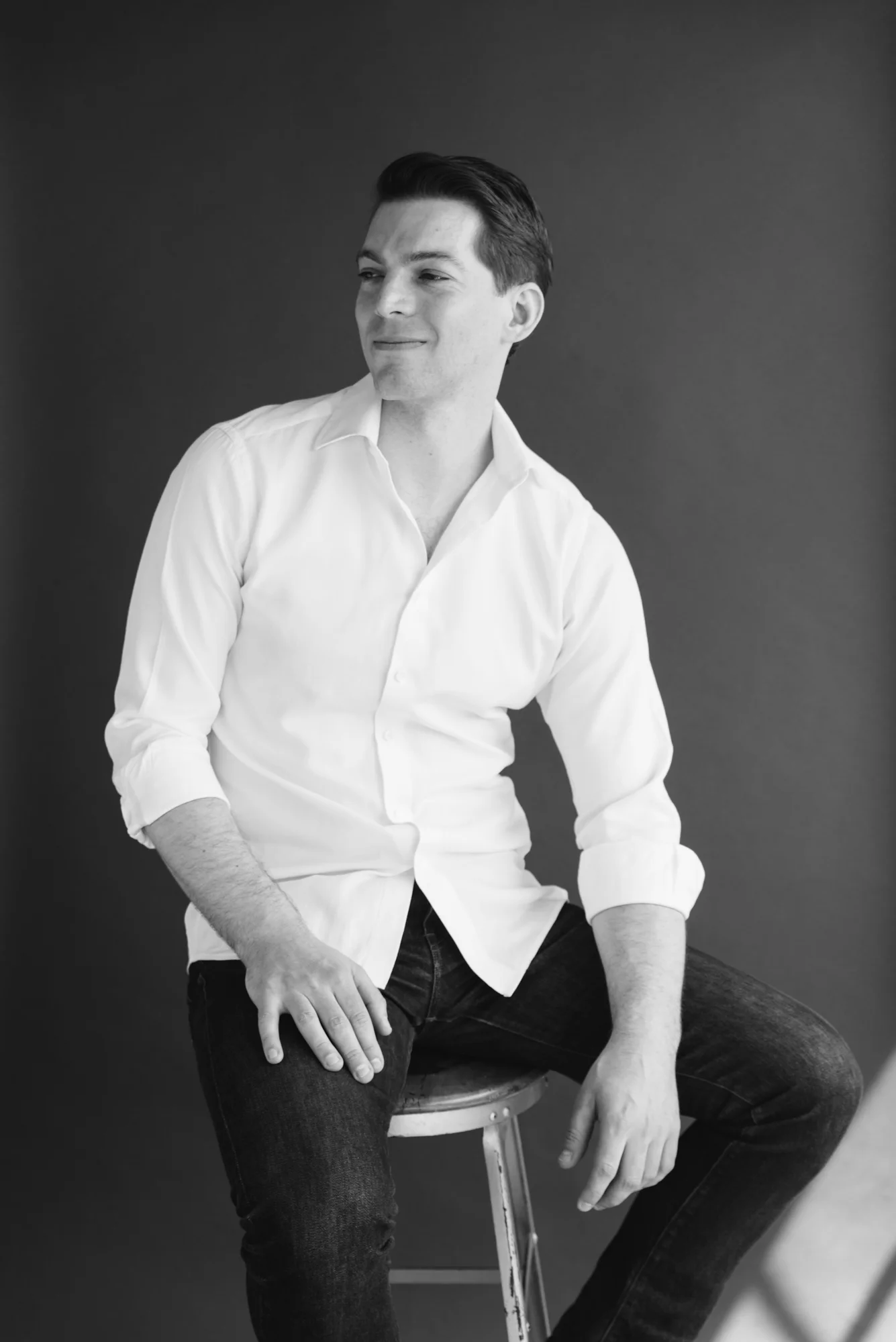 Black and white studio headshot of a man sitting on a stool in a white button up shirt and dark jeans, looking off to the side