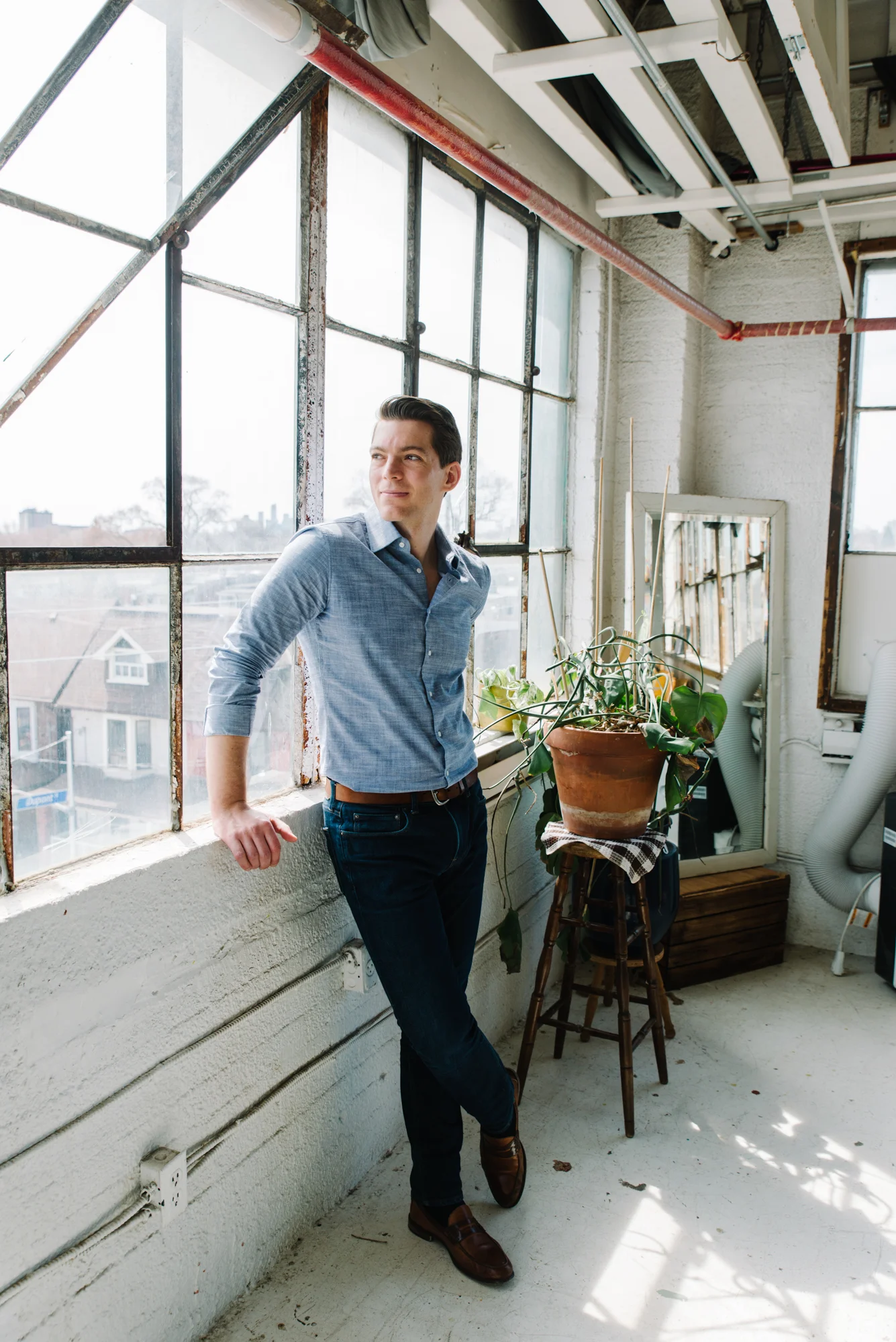 Casual studio portrait of a man leaning against large bright industrial windows in a blue button up shirt and jeans