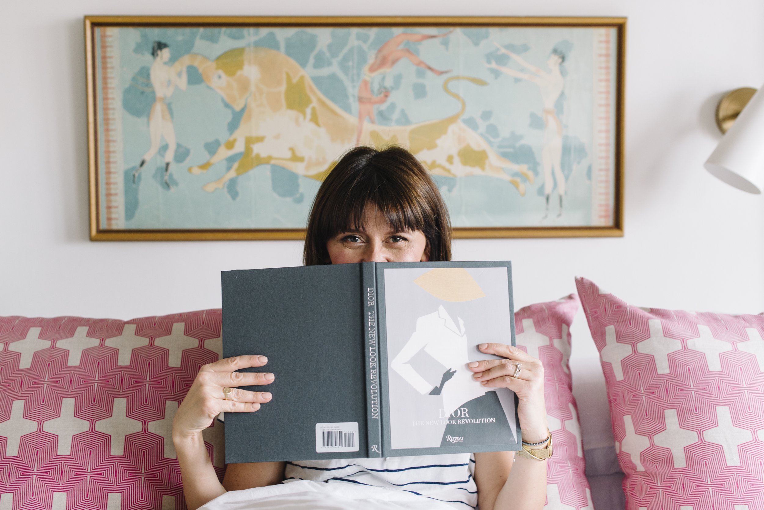 Playful branding portrait of interior designer Rebecca Hay seated on a couch with pink cushions, holding a book up with her eyes peeking over the top