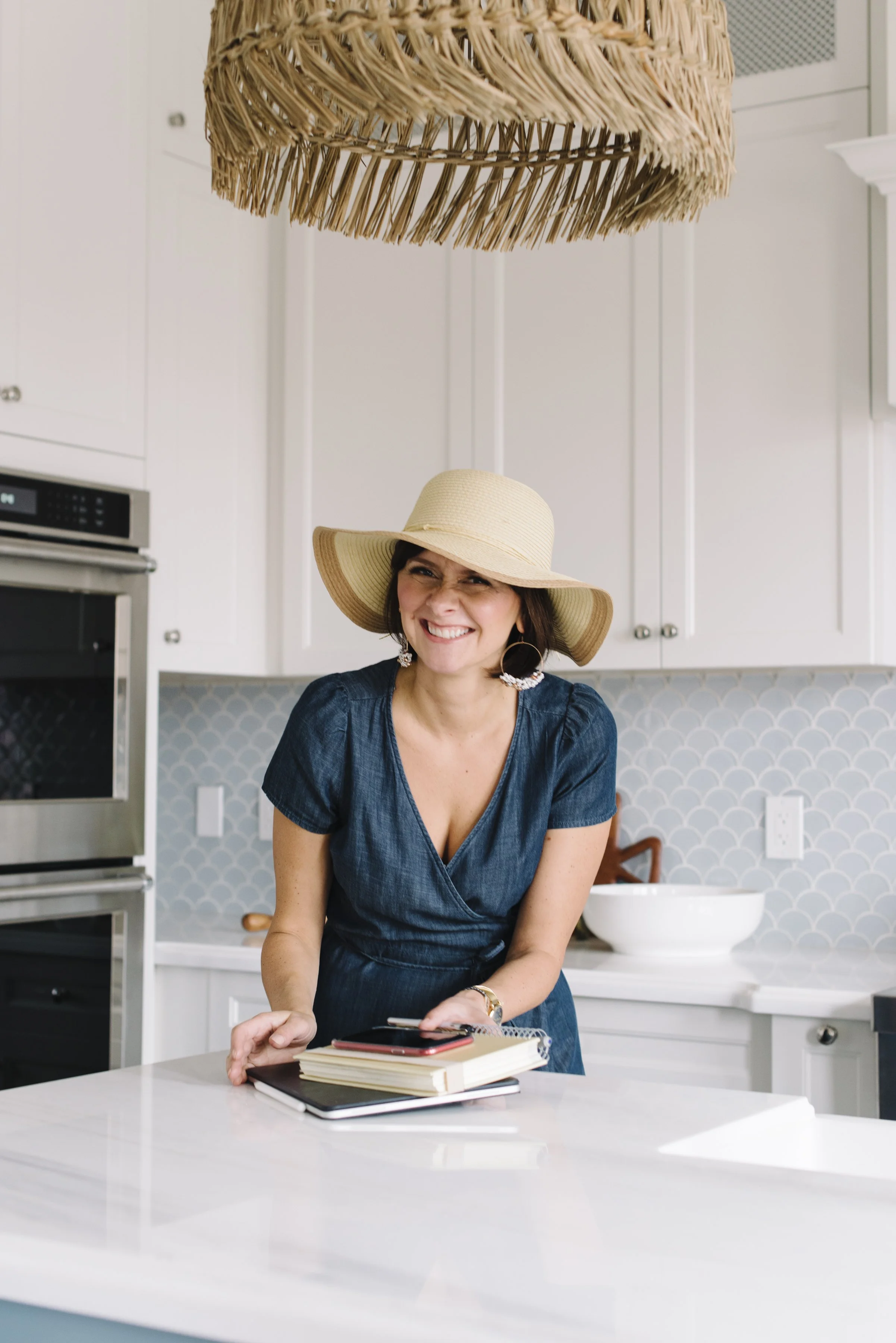 Lifestyle branding photo of Rebecca Hay at a kitchen island grabbing her laptop and phone, wearing a wide brimmed hat and smiling at the camera