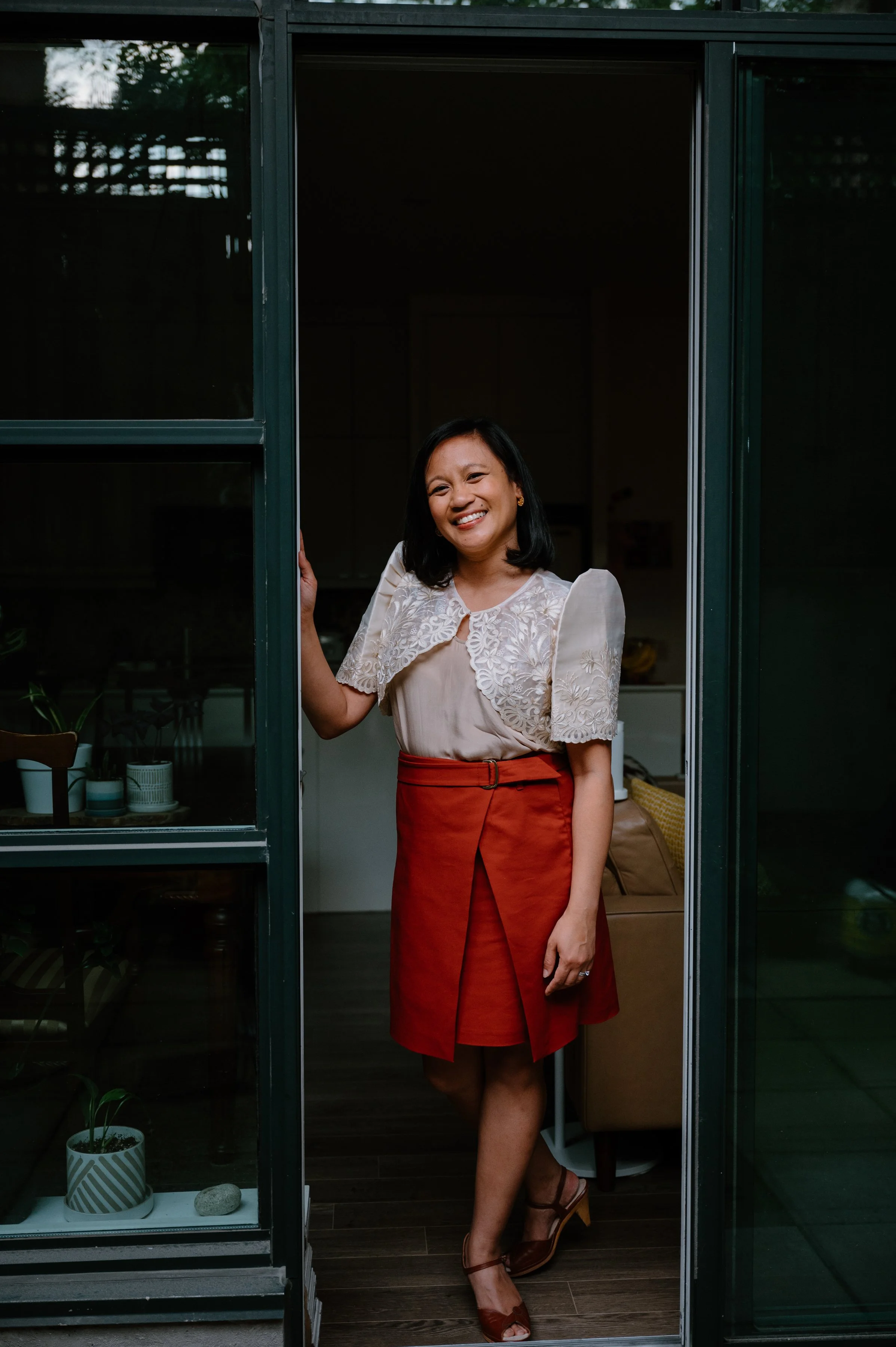 Editorial portrait of Jennilee Austria-Bonifacio standing in her open doorway wearing a Filipiniana terno, smiling at the camera