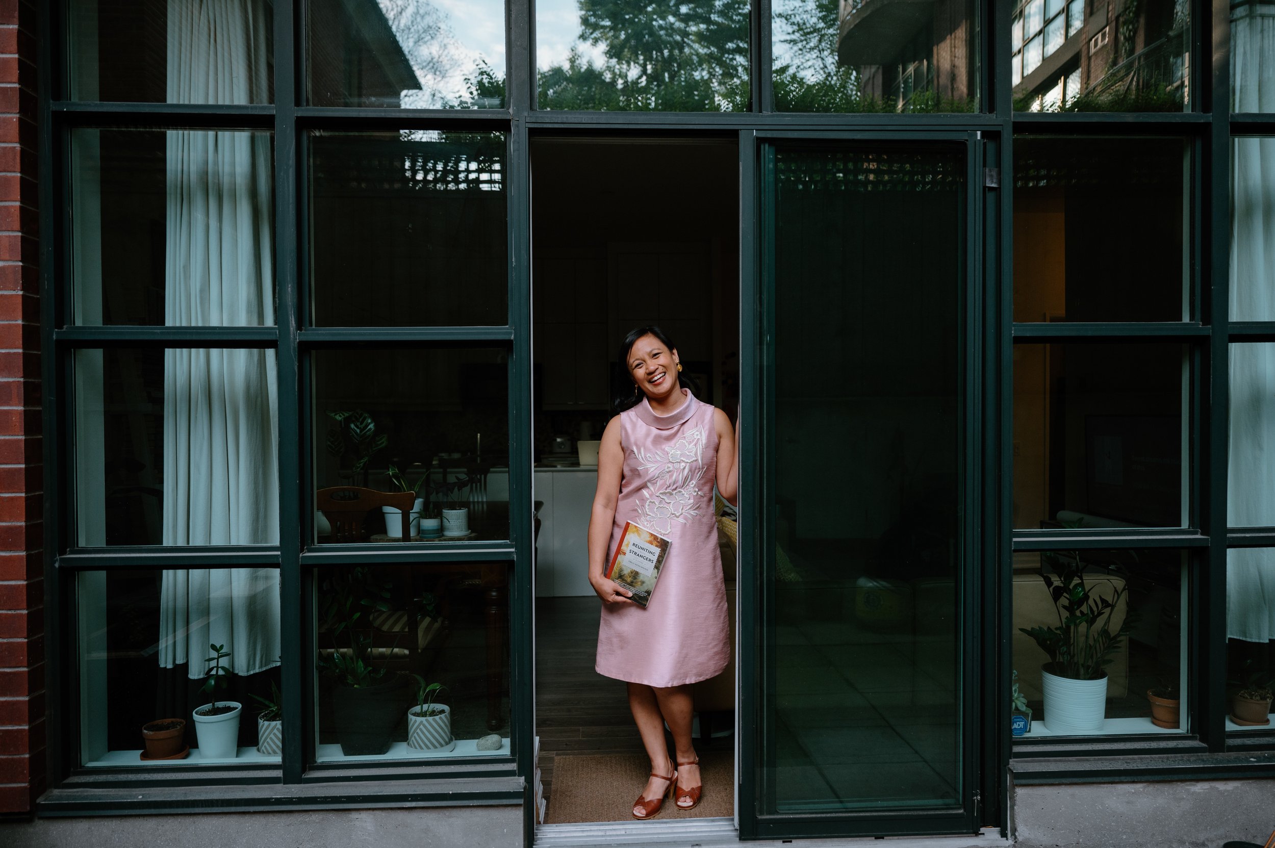 Lifestyle author portrait of Jennilee Austria-Bonifacio standing in her open doorway at home in Toronto, holding a copy of Reuniting with Strangers