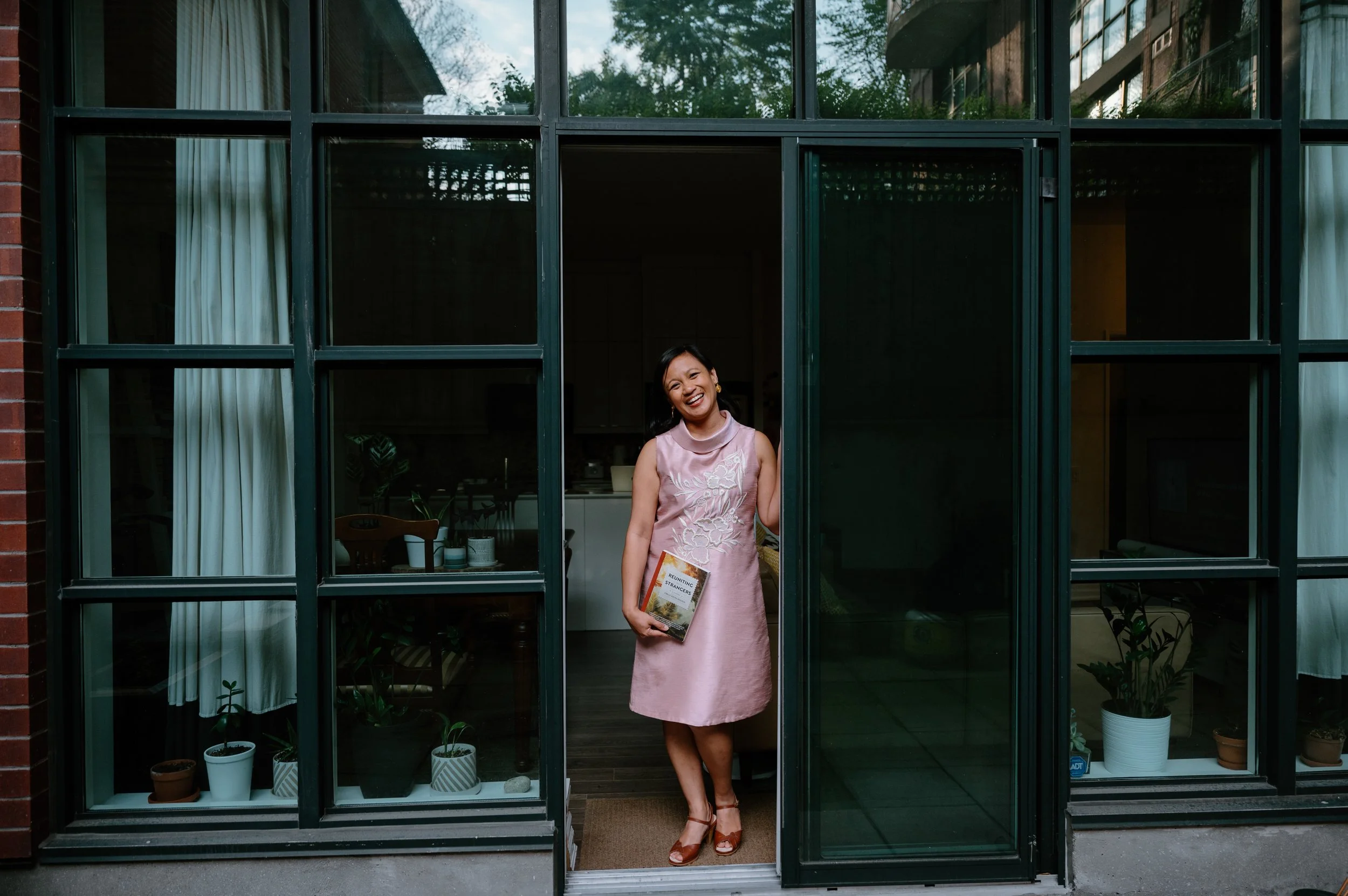 Editorial portrait of author Jennilee Austria-Bonifacio standing in her open doorway holding her debut novel Reuniting with Strangers