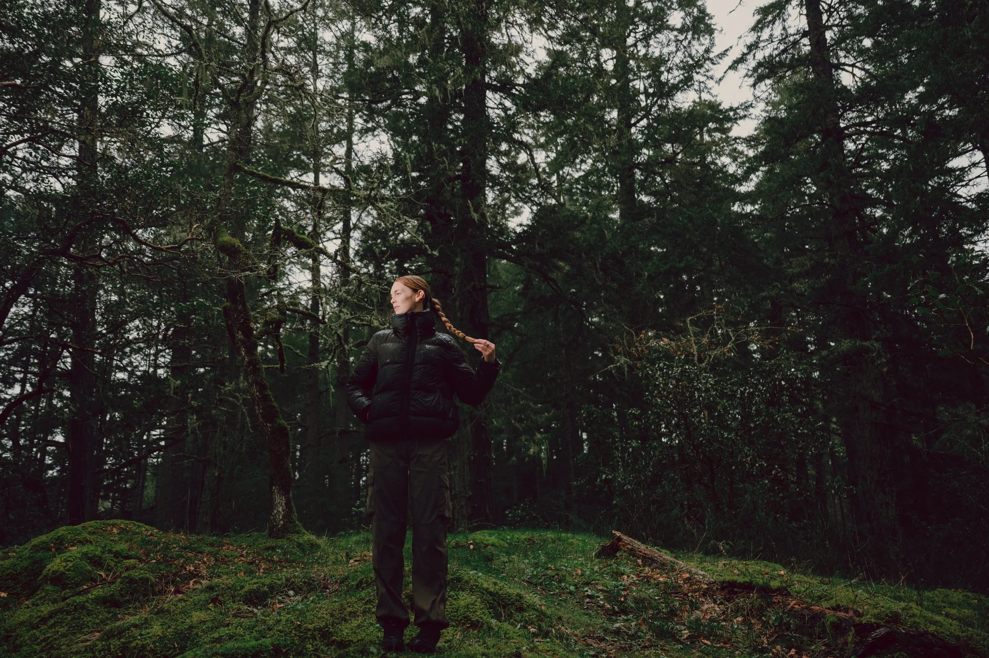 Wide gorpcore-inspired campaign photo of a model standing on a moss-covered hill at Thetis Lake surrounded by BC forest