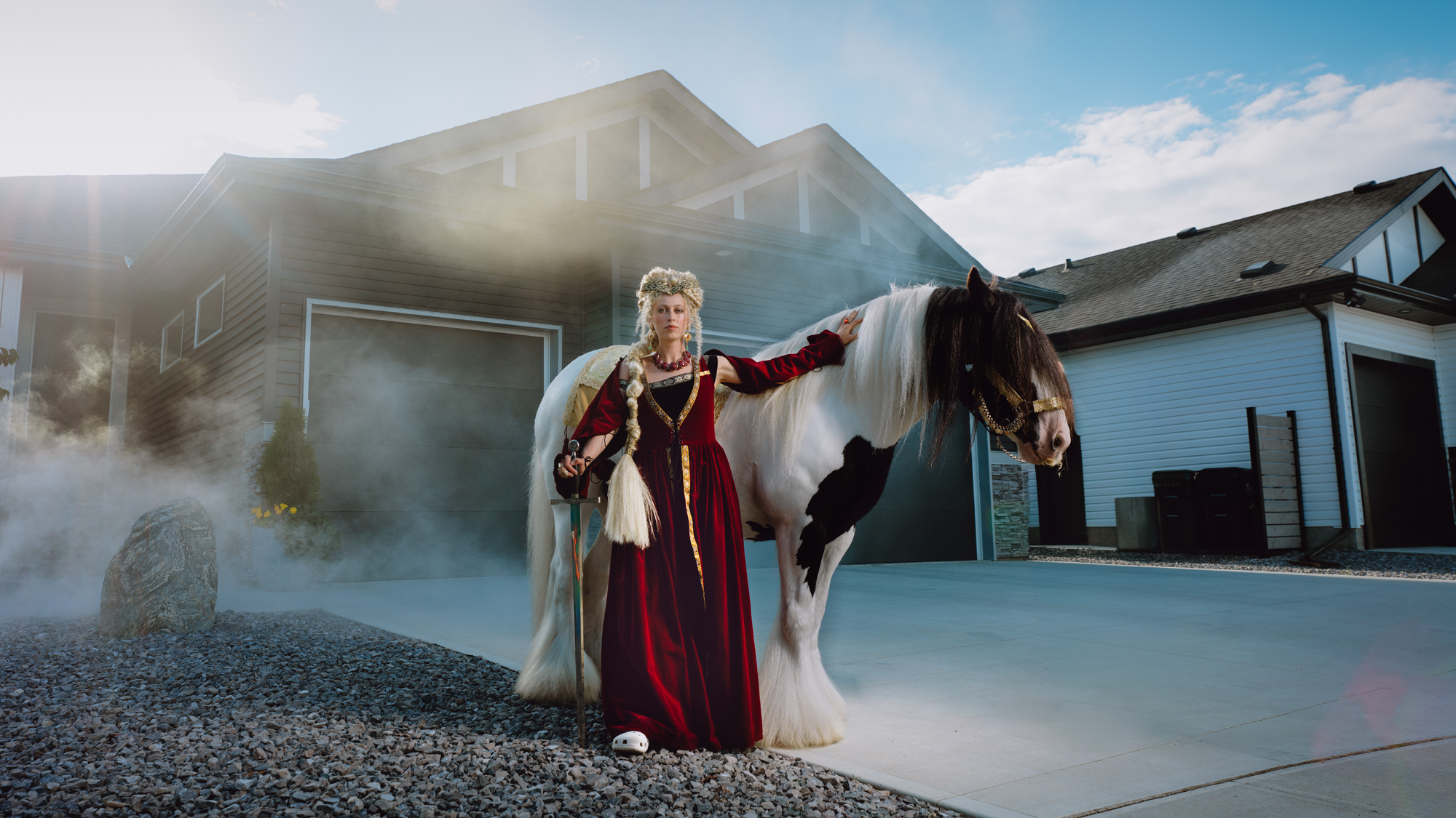 A woman dressed in medieval-style red and black gown with a long braid, standing next to a black and white horse in front of a modern house with a garage, with smoke or mist in the background and a partly cloudy sky.