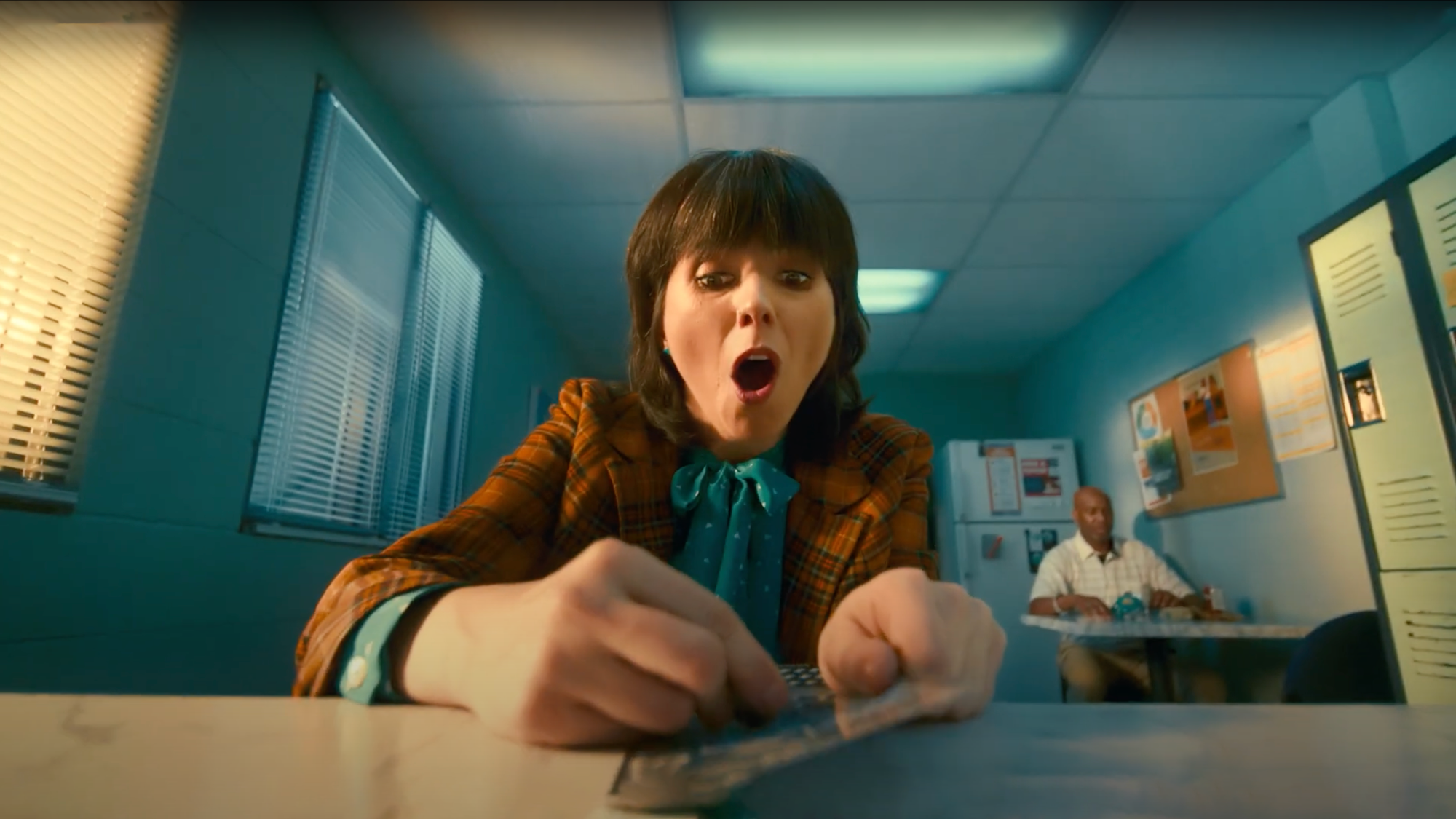 A woman with dark hair and a plaid blazer looks shocked or surprised while handling a small object on a table in a brightly lit office break room. In the background, a man is sitting at a table.