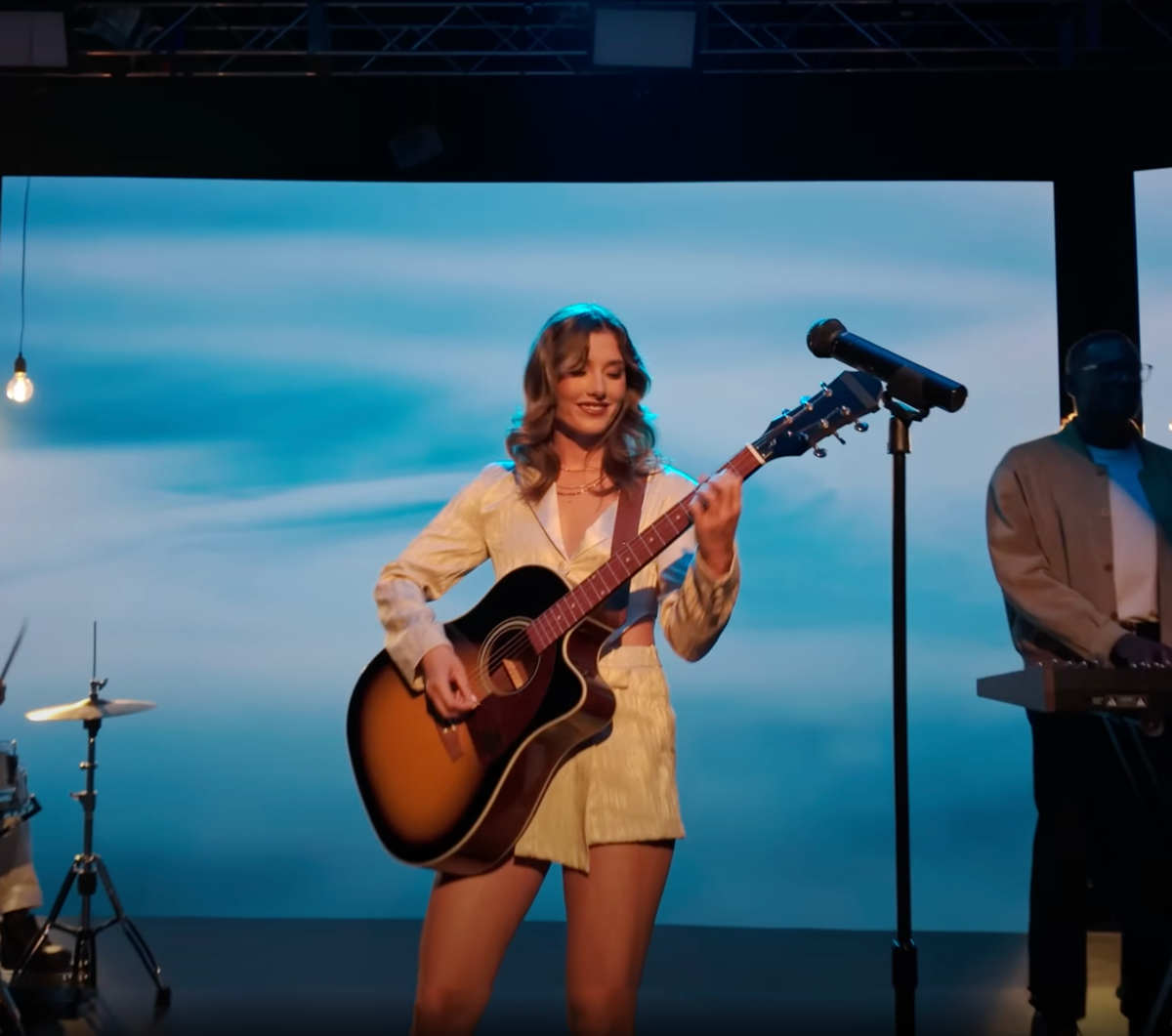 Woman playing an acoustic guitar on stage during a live music performance, with a blue sky background on a large screen behind her.