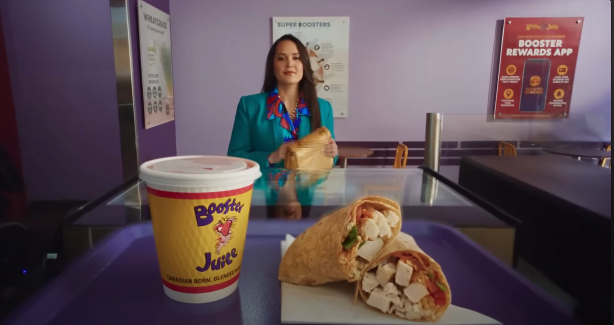 A woman in a teal blazer holding a sandwich at a food counter. In the foreground, a cup of Booster Juice and a wrap sandwich filled with chicken, vegetables, and cheese on a purple tray.