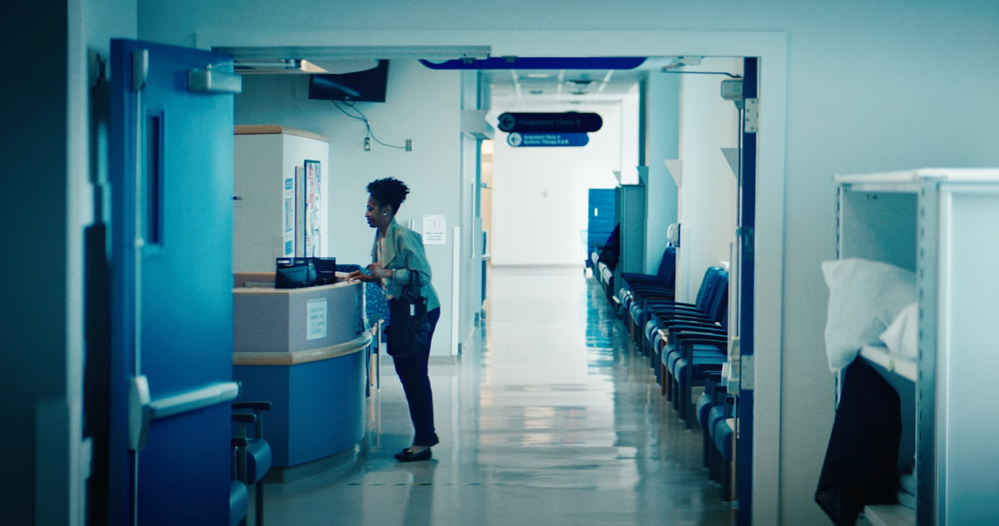 A woman at a hospital reception desk in a corridor with chairs and hospital beds.
