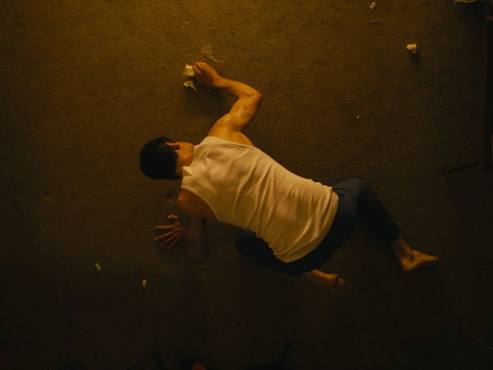 A person with dark hair in a white sleeveless shirt and dark pants is lying on their side on a brown carpeted floor, appearing to be resting or injured, with some pieces of white tissue or paper near them.