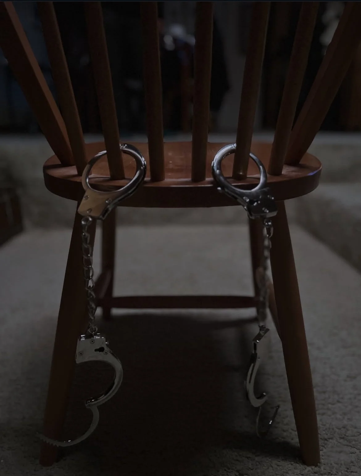 A wooden chair with jail handcuffs hanging from its top rail, positioned in a dimly lit room.