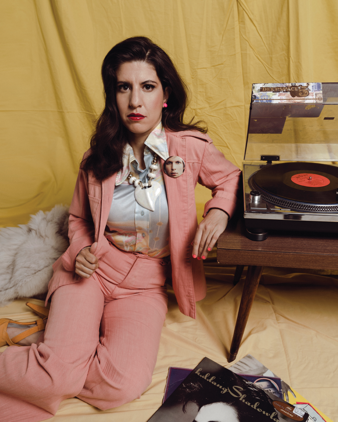 A woman with dark wavy hair, wearing a pink suit and colorful blouse, sits on the floor next to a turntable with a record. She has a large heart-shaped necklace and a pin with a face on her blazer. The background is yellow, with magazines and records on the floor.