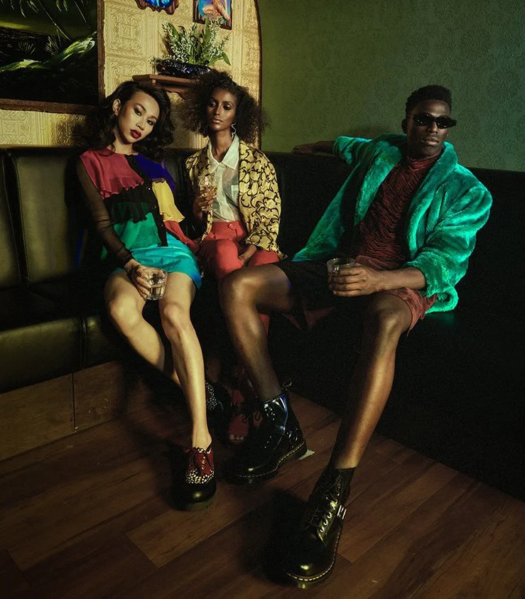 Three young adults sitting on a retro-style green and black couch in a dimly lit room, dressed in colorful fashion, with drinks in hand, showcasing a vibrant and stylish scene.