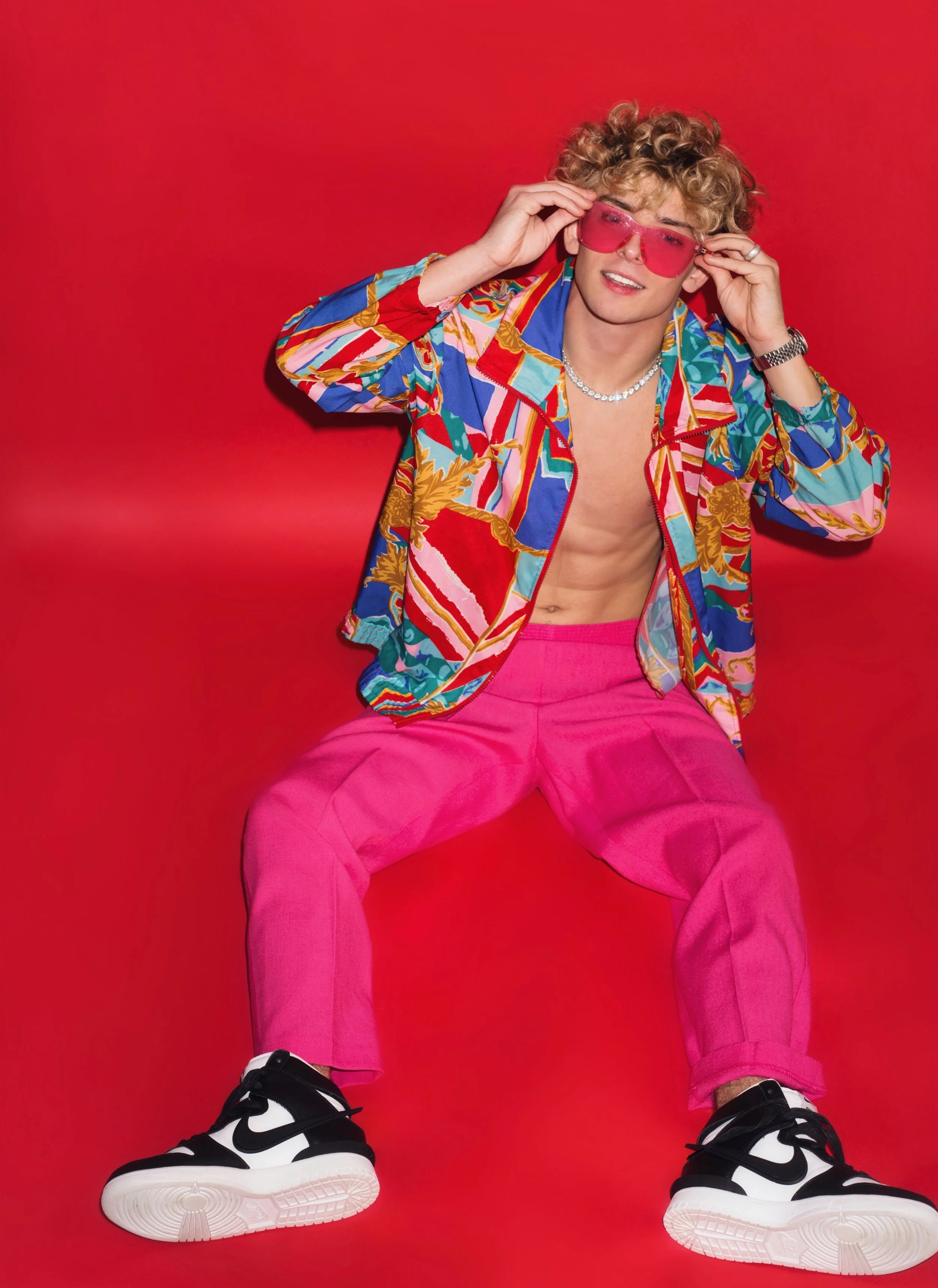Person with curly hair wearing pink sunglasses, a colorful patterned jacket, pink pants, and black and white sneakers, sitting against a red background.