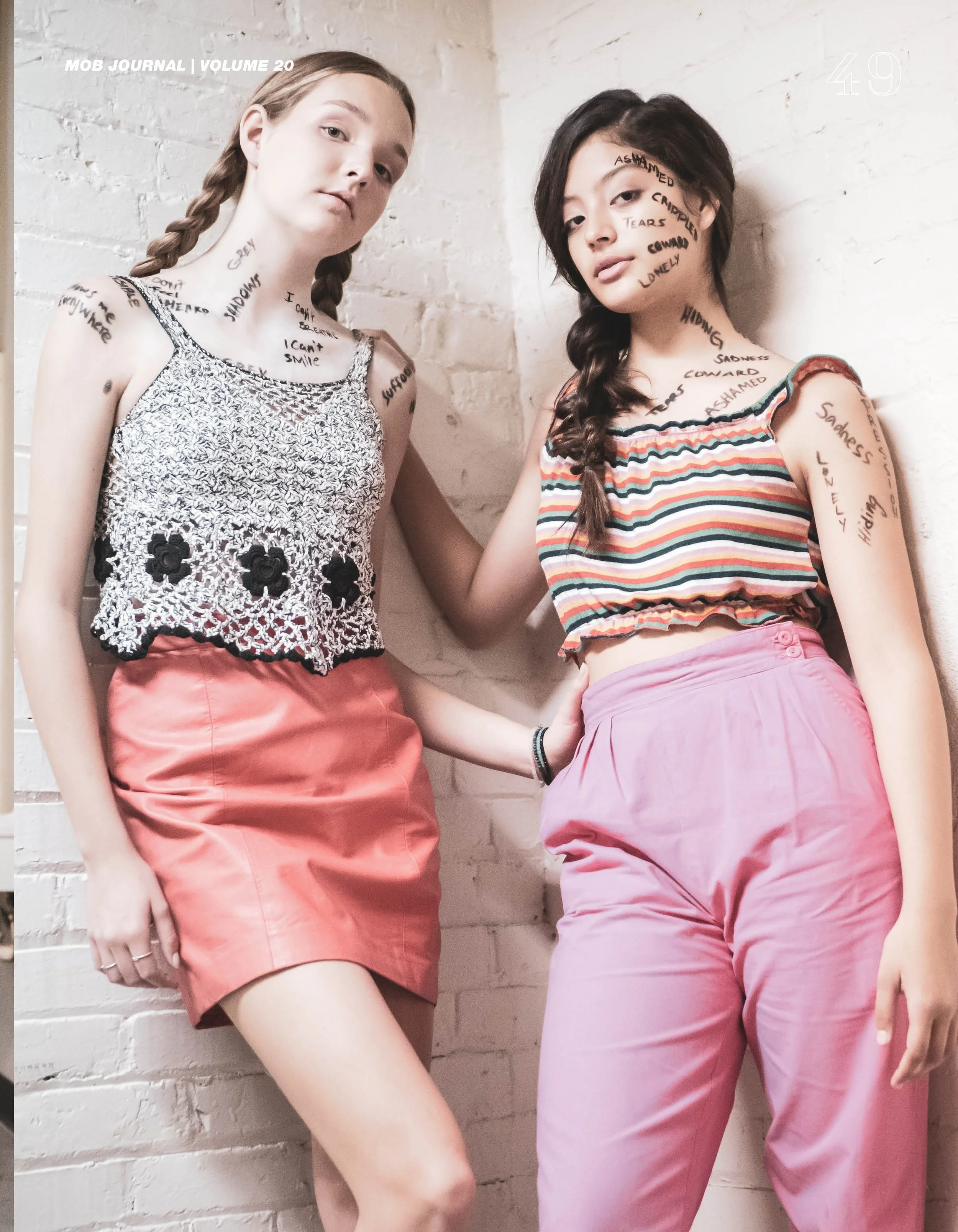 Two young women with text written on their faces and arms, leaning against a white brick wall, dressed in colorful casual clothing, with one wearing a sleeveless top with lace and floral design and a pink skirt, and the other in a striped tank top with pink pants.
