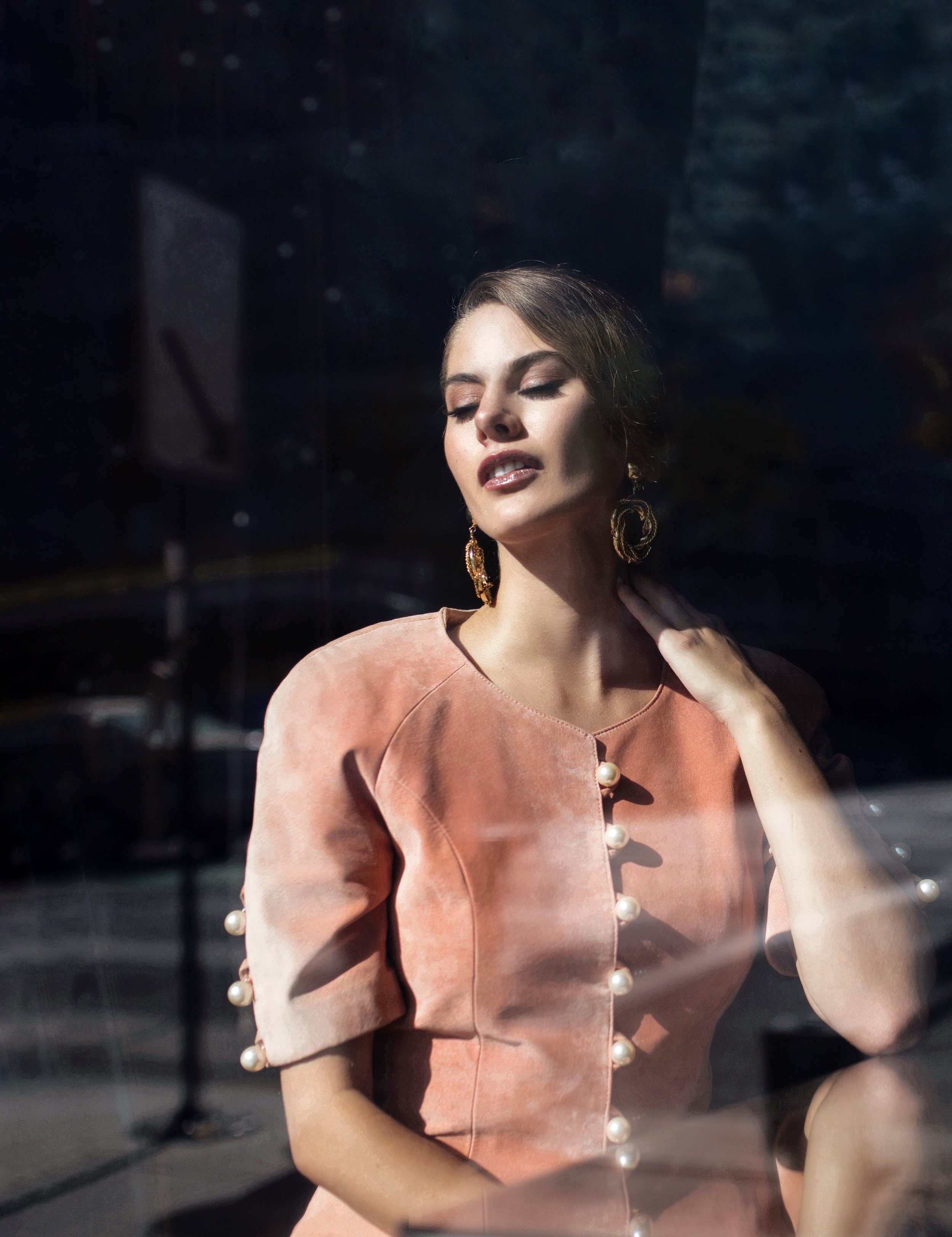 A woman with closed eyes and slightly parted lips, touching her neck, standing behind a glass window. She is wearing a peach-colored dress with pearl button details and large gold earrings. The sunlight highlights her face and upper body.