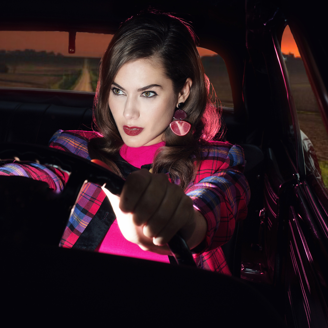 Woman with dark wavy hair wearing a plaid jacket and pink turtleneck, driving a car at sunset.