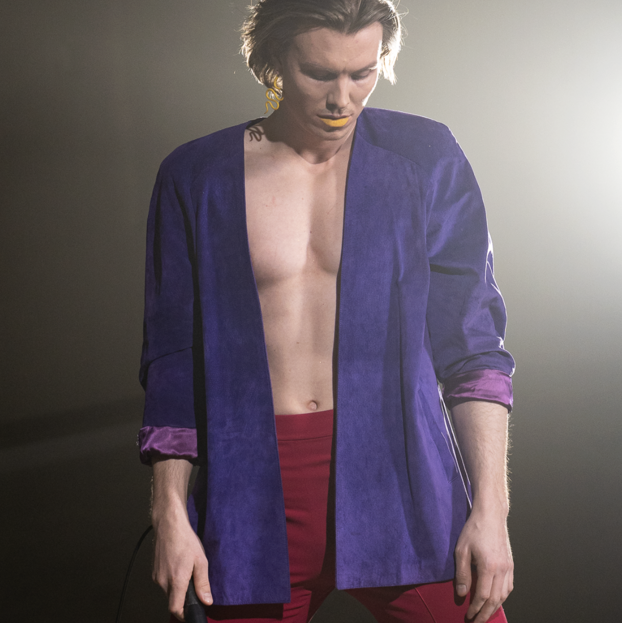 A male performer with shoulder-length hair, yellow lipstick, and dramatic eye makeup, standing on stage with a microphone in hand. He is wearing a purple velvet blazer with rolled-up sleeves, revealing pink lining, and red pants. The background is da