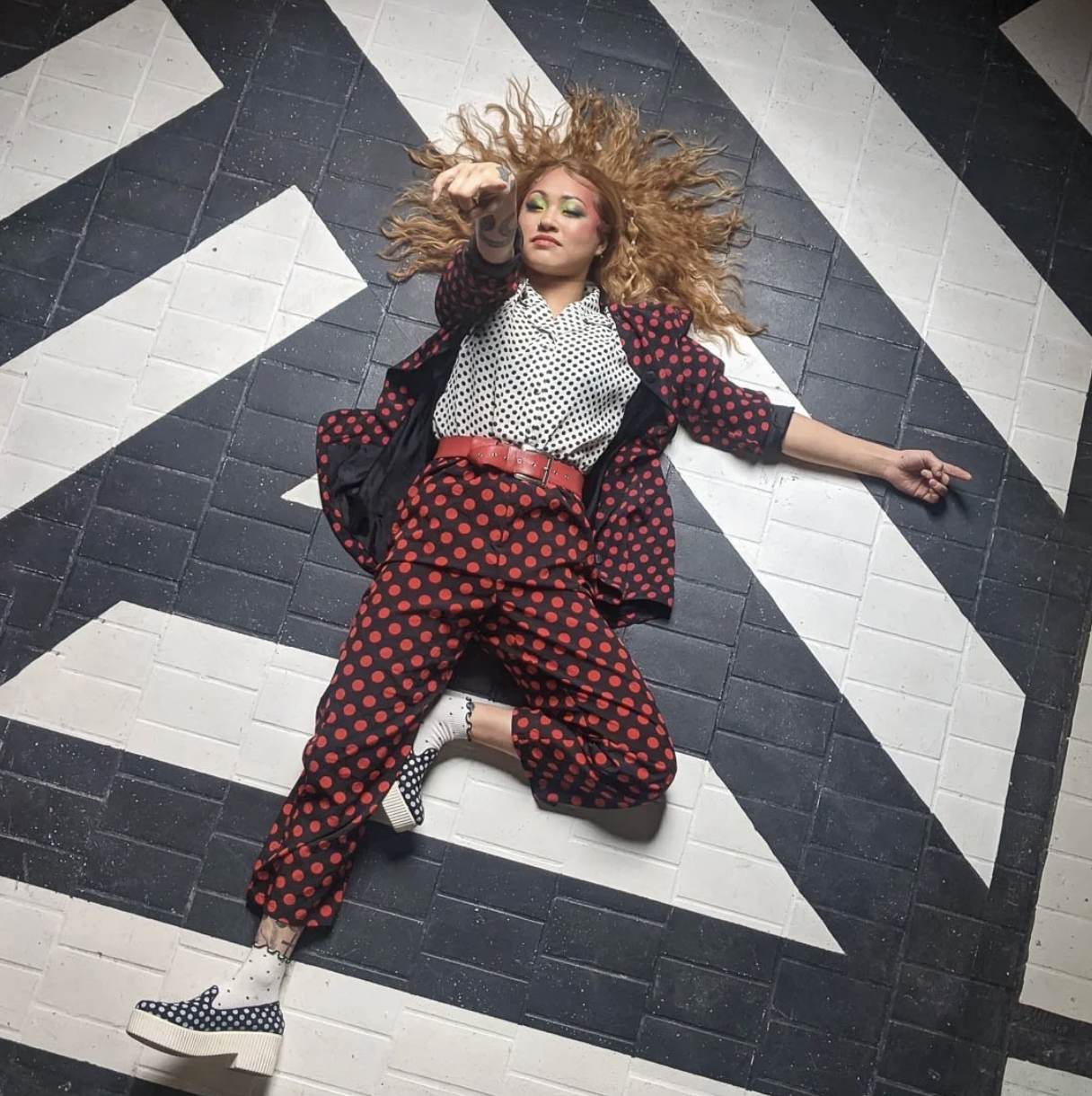 Woman with curly hair lying on a geometric black and white tiled floor, wearing a polka-dot outfit and colorful makeup.