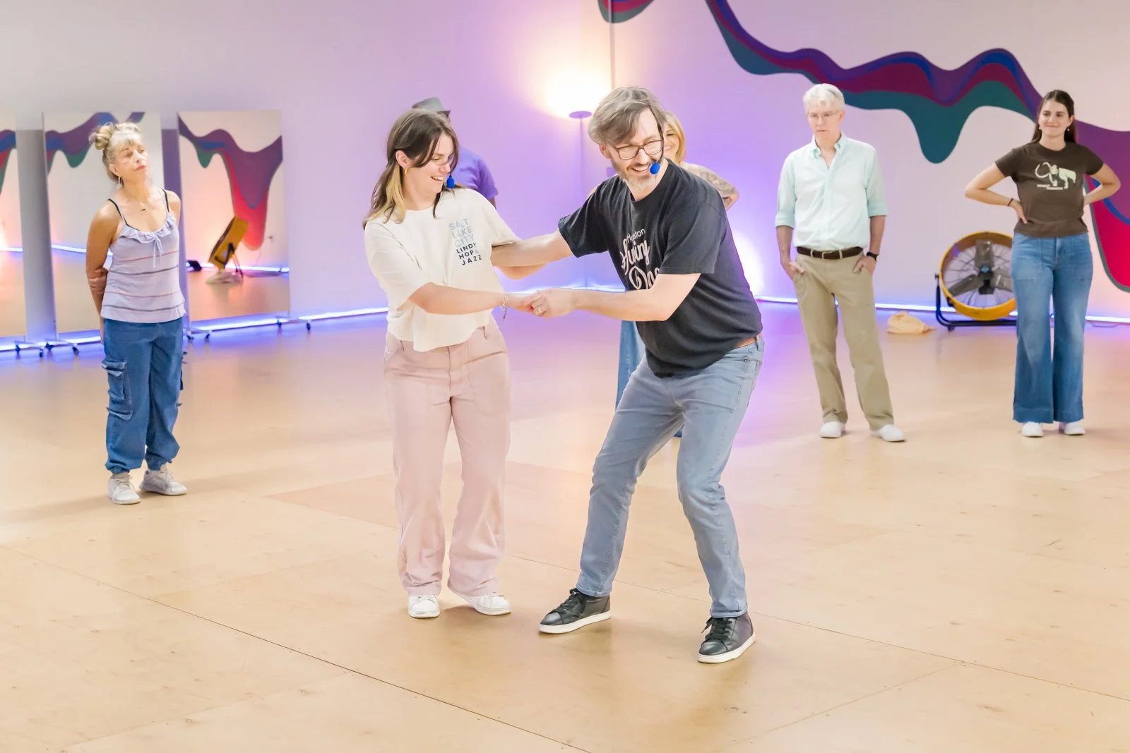 A group of people in a dance space with colorful abstract wall art. Two people in the front are dancing and smiling, holding hands. Other individuals are standing in the background, watching or waiting for their turn.
